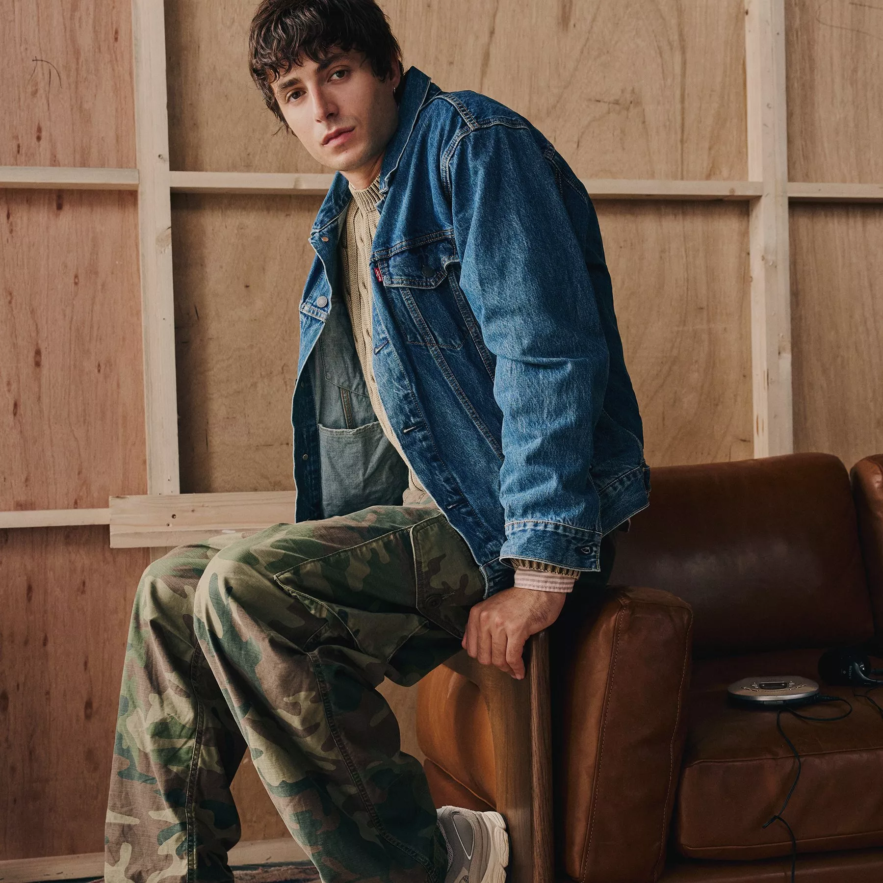 A man sits on a leather sofa wearing a denim jacket, cable-knit sweater, and camouflage pants in front of a wooden wall