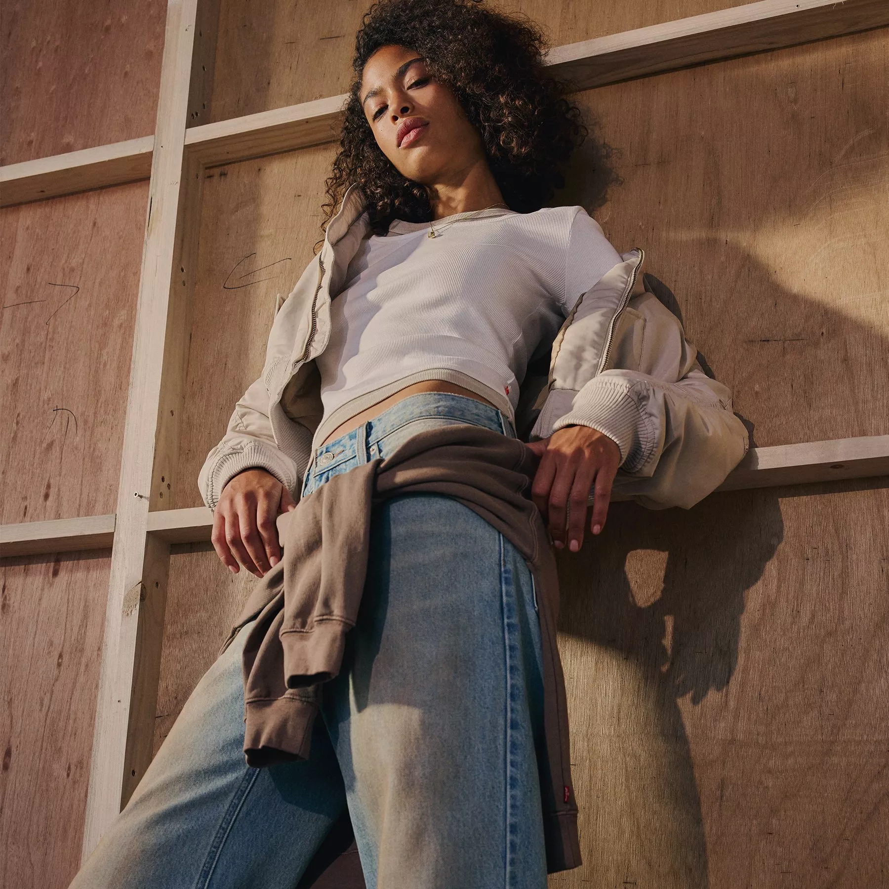 A woman with curly hair leans against a wooden wall, wearing a white ribbed top, light-wash jeans, and a neutral-toned jacket