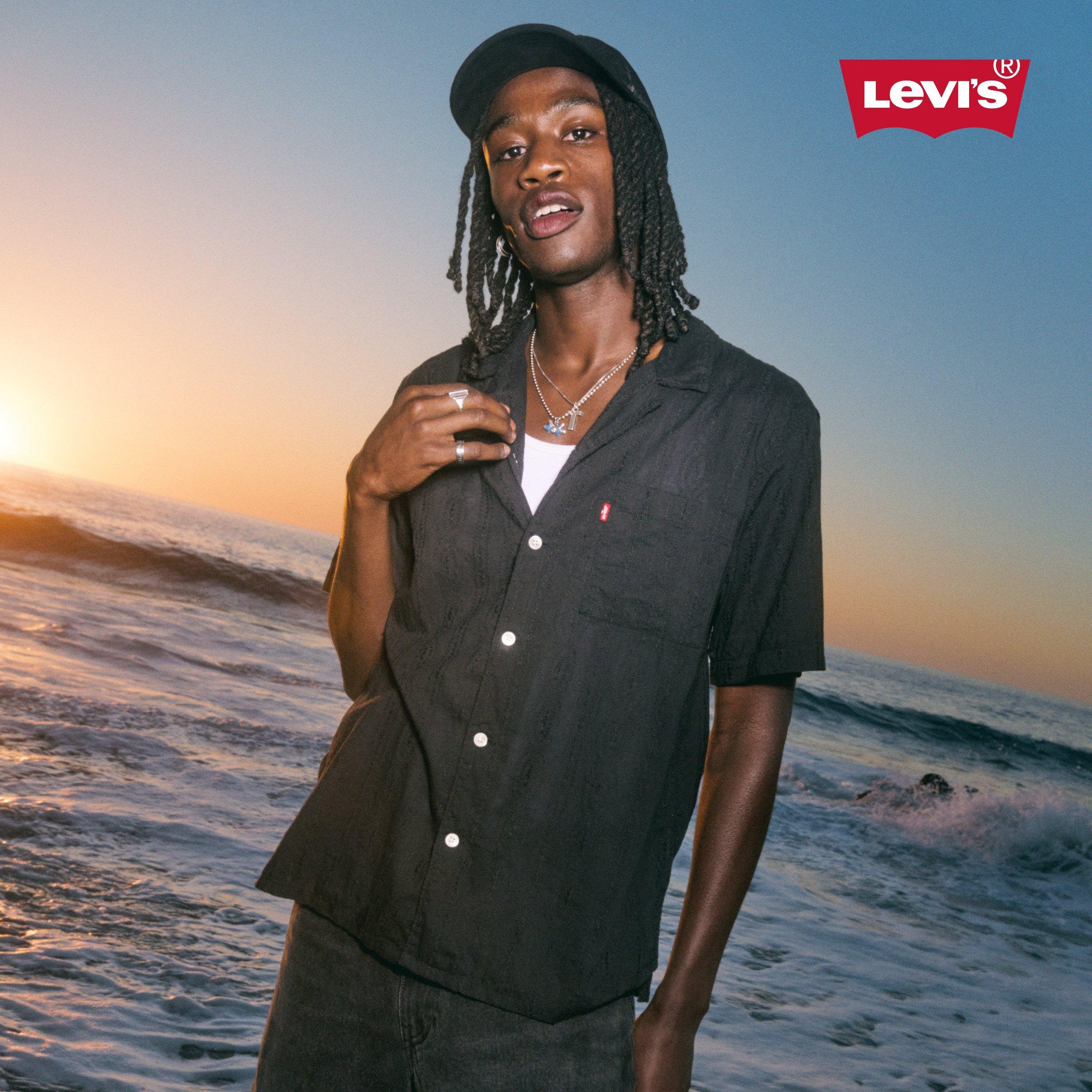 A person standing by the ocean at sunset, wearing a black shirt, cap, and jewelry, with the Levi's logo in the top right corner.
