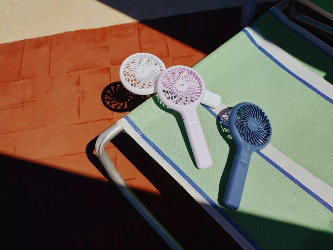 Three portable handheld mini fans on a green striped sun lounger, casting shadows on terracotta tiles outdoors