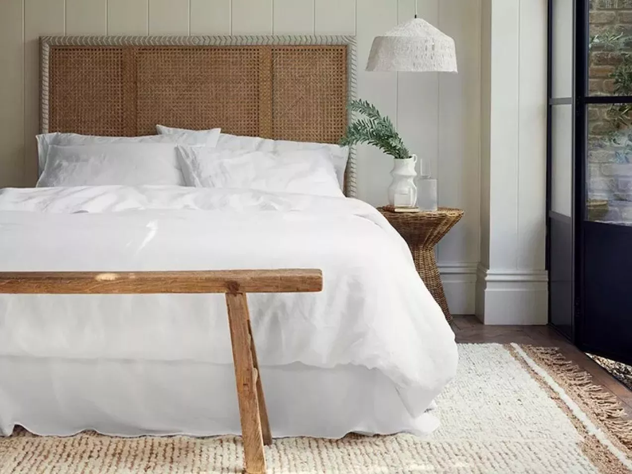 Bright coastal bedroom with white bedding, rattan headboard, woven side table, pendant light and rug by window
