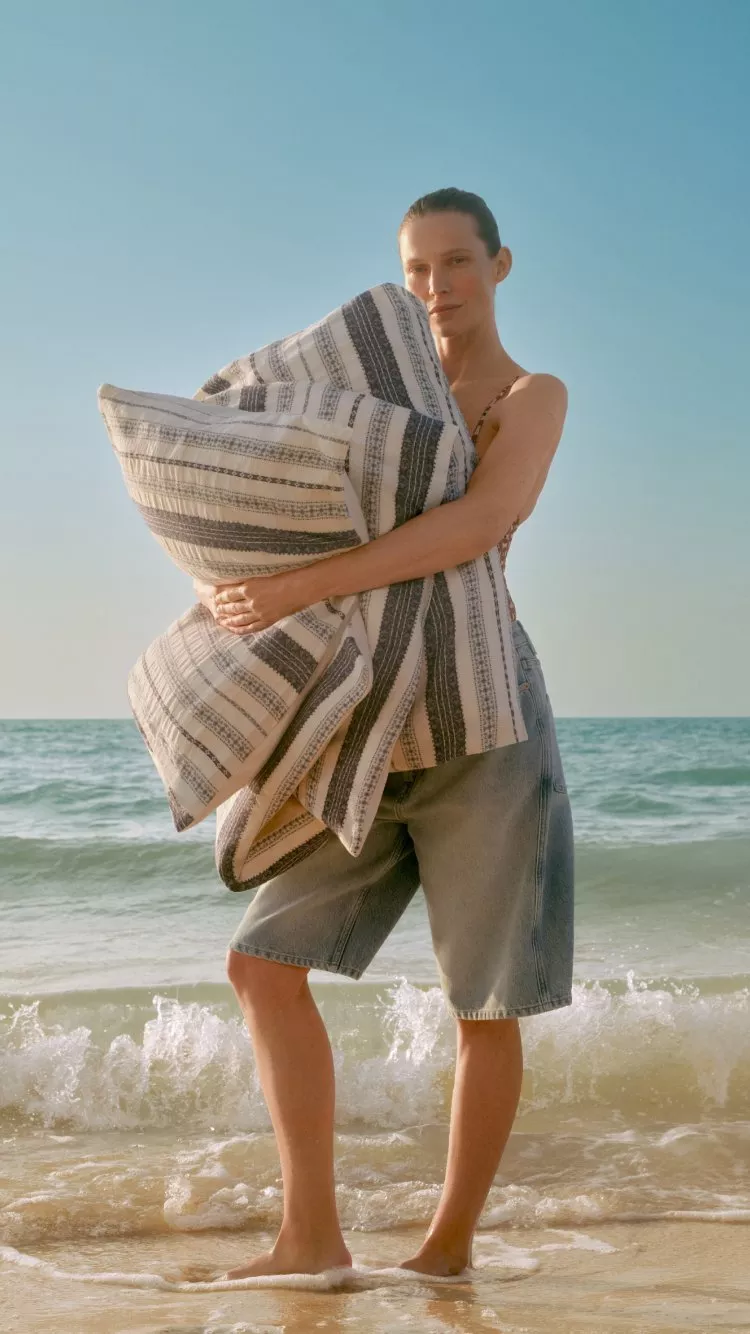 Woman on a sunny beach holding a large striped cushion, wearing denim shorts with waves and blue sky behind