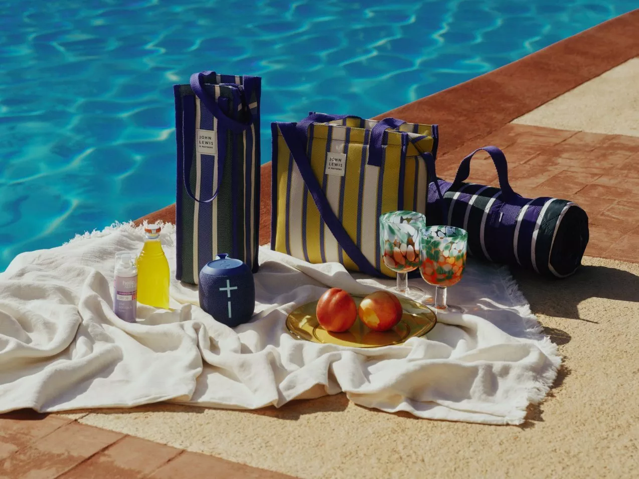 Poolside patio with a picnic with striped tote bags, blanket, fruit and drinks, beside a sunlit swimming pool