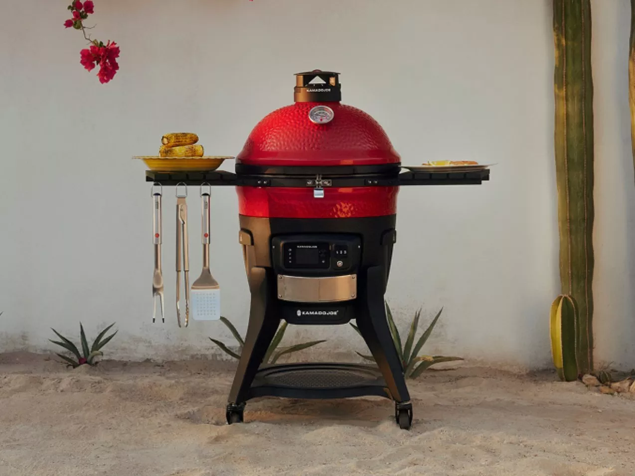 Red kamado charcoal barbecue grill with side shelves, utensils and corn, set in an outdoor scene.