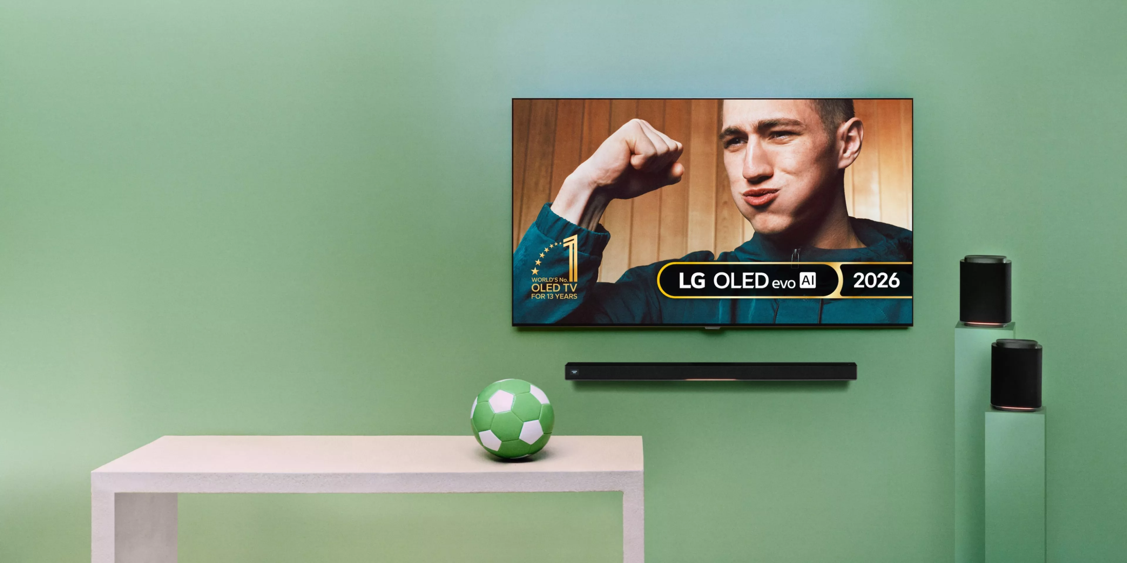 A table with a green football on it, beside an LG OLED evo AI 2026 TV on green wall with soundbar, showing a man celebrating 