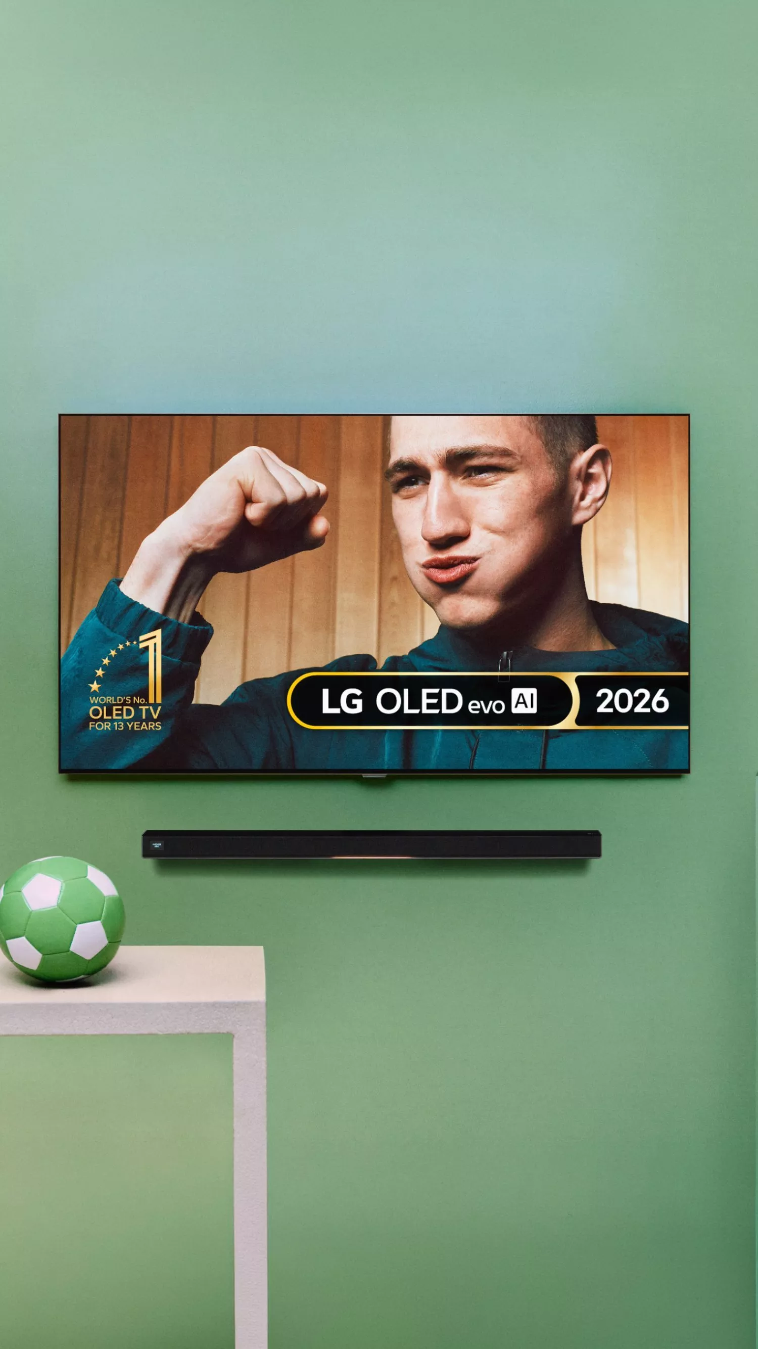 A table with a green football on it, beside an LG OLED evo AI 2026 TV on green wall with soundbar, showing a man celebrating 