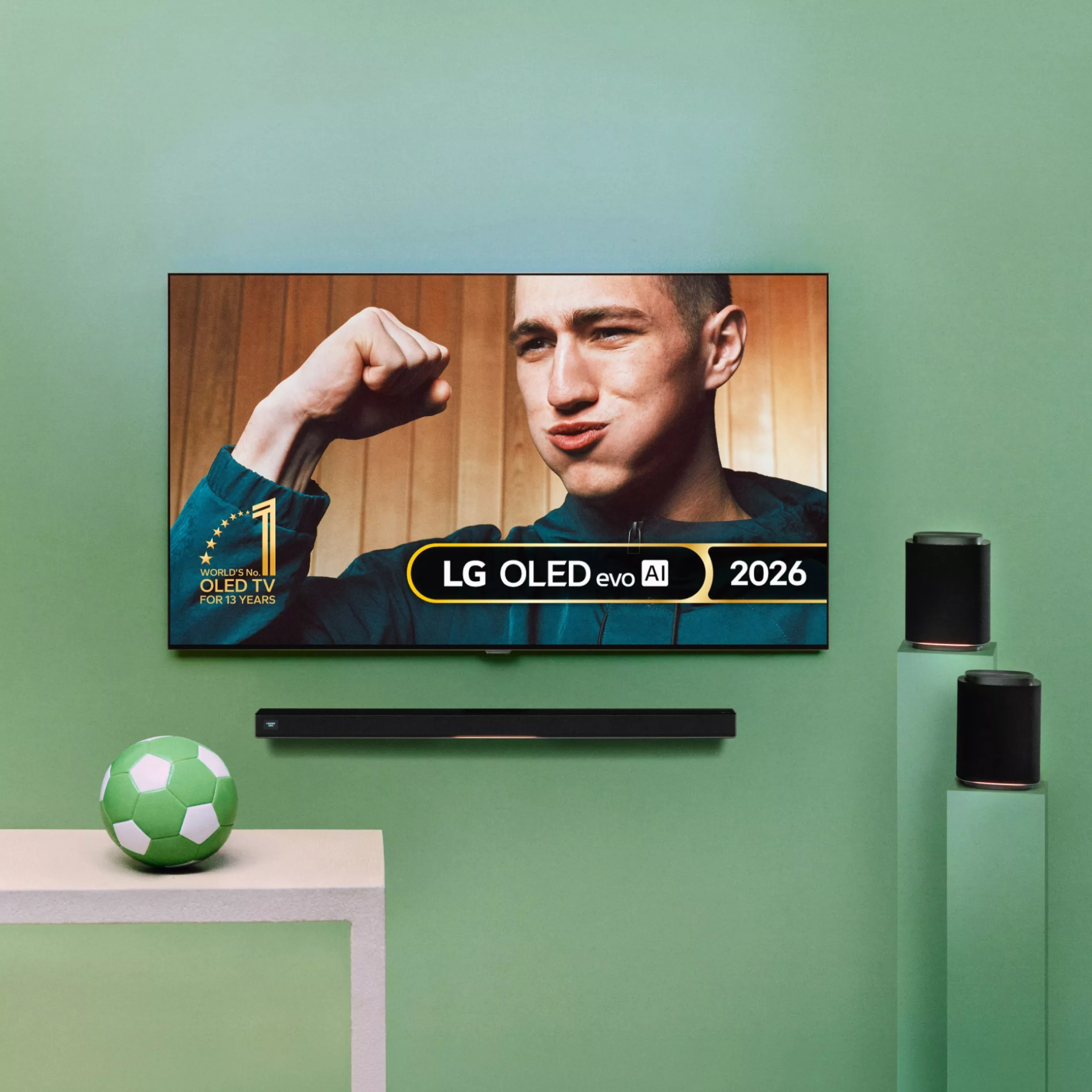 LG OLED evo AI 2026 TV on green wall with soundbar and speakers, showing man flexing on screen in living room