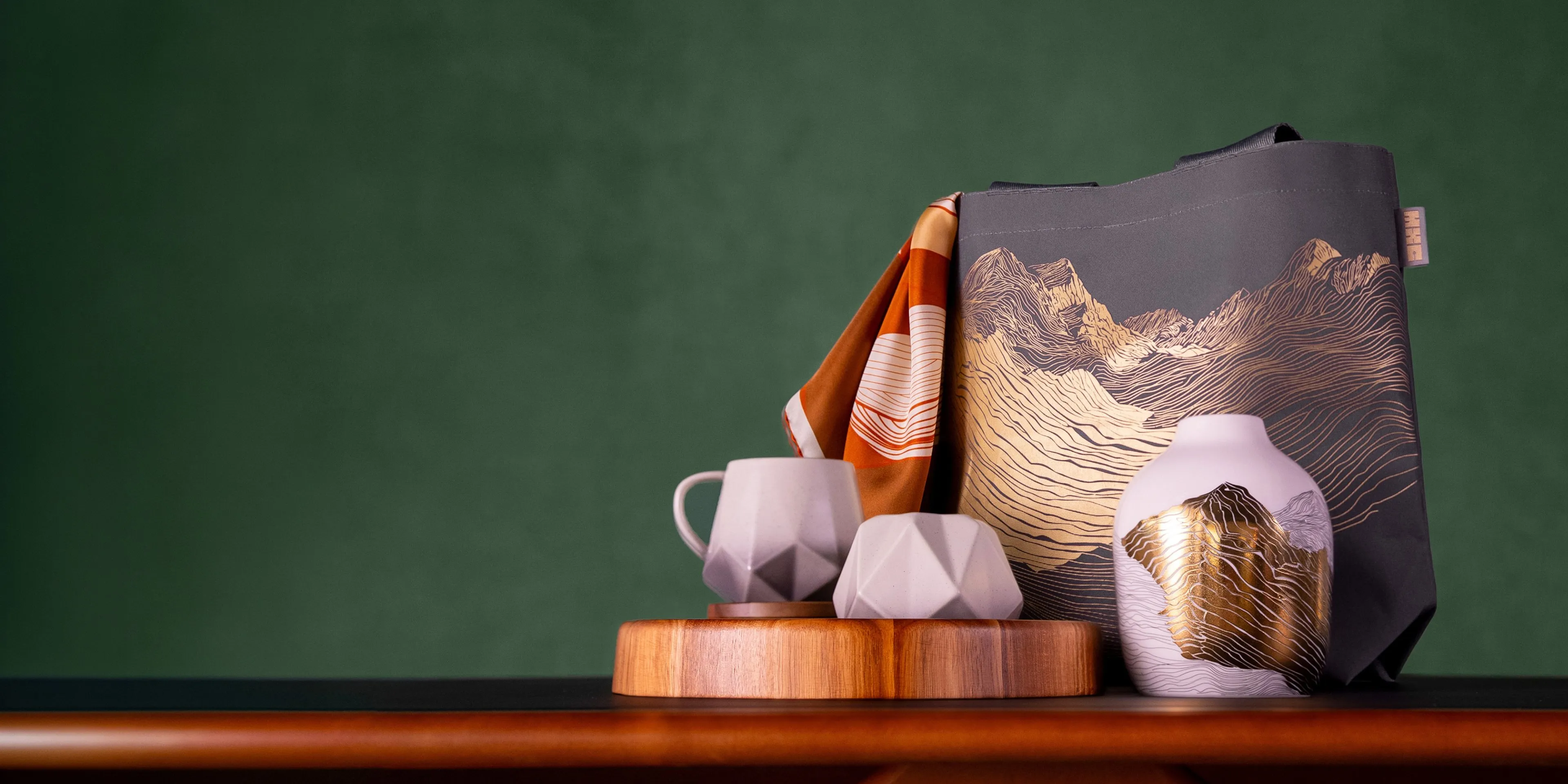 Decorative ceramic mug, bowl and vase with mountain print, plus tote bag and scarf on wooden board, green backdrop