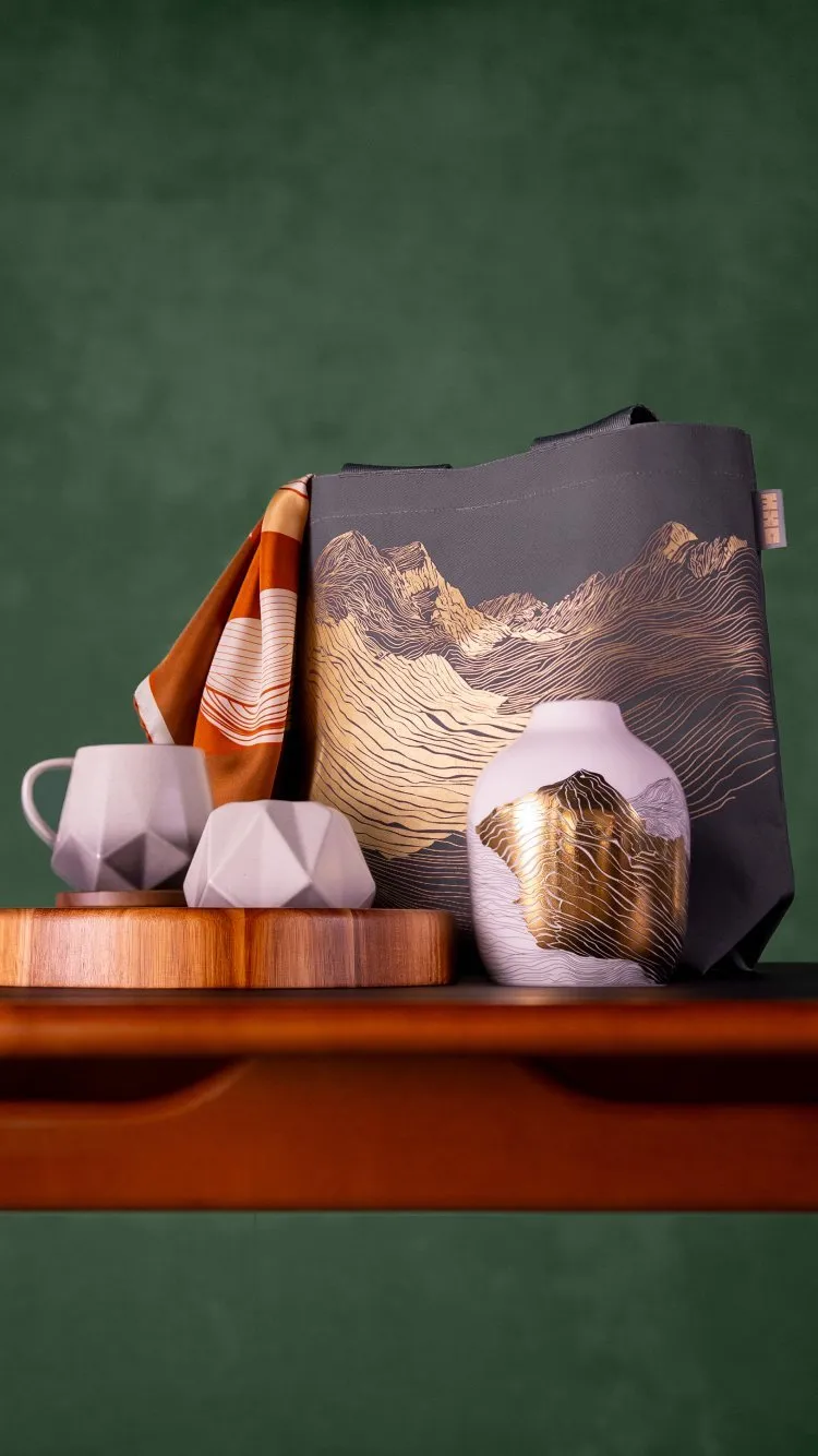Grey tote bag with mountain line art, scarf, ceramic mugs and vase on wooden tray against green wall