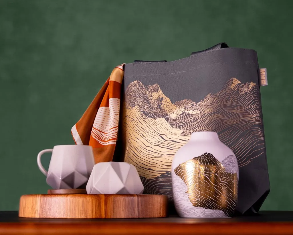 Grey tote bag with gold mountain line art, ceramic mug and vase on wooden tray, against a green background
