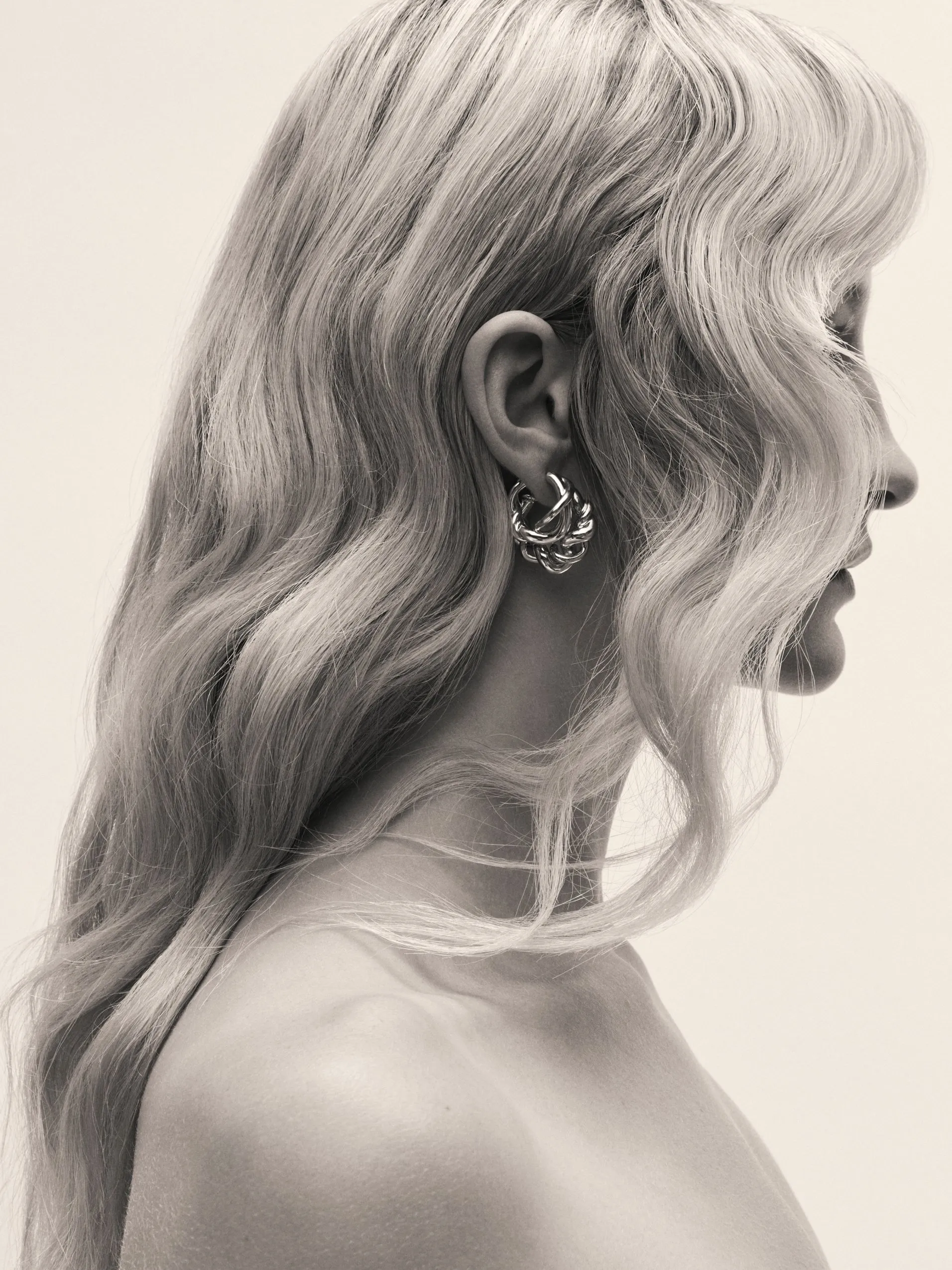 Black and white side profile portrait of blonde woman with wavy hair wearing chunky hoop earrings in studio lighting