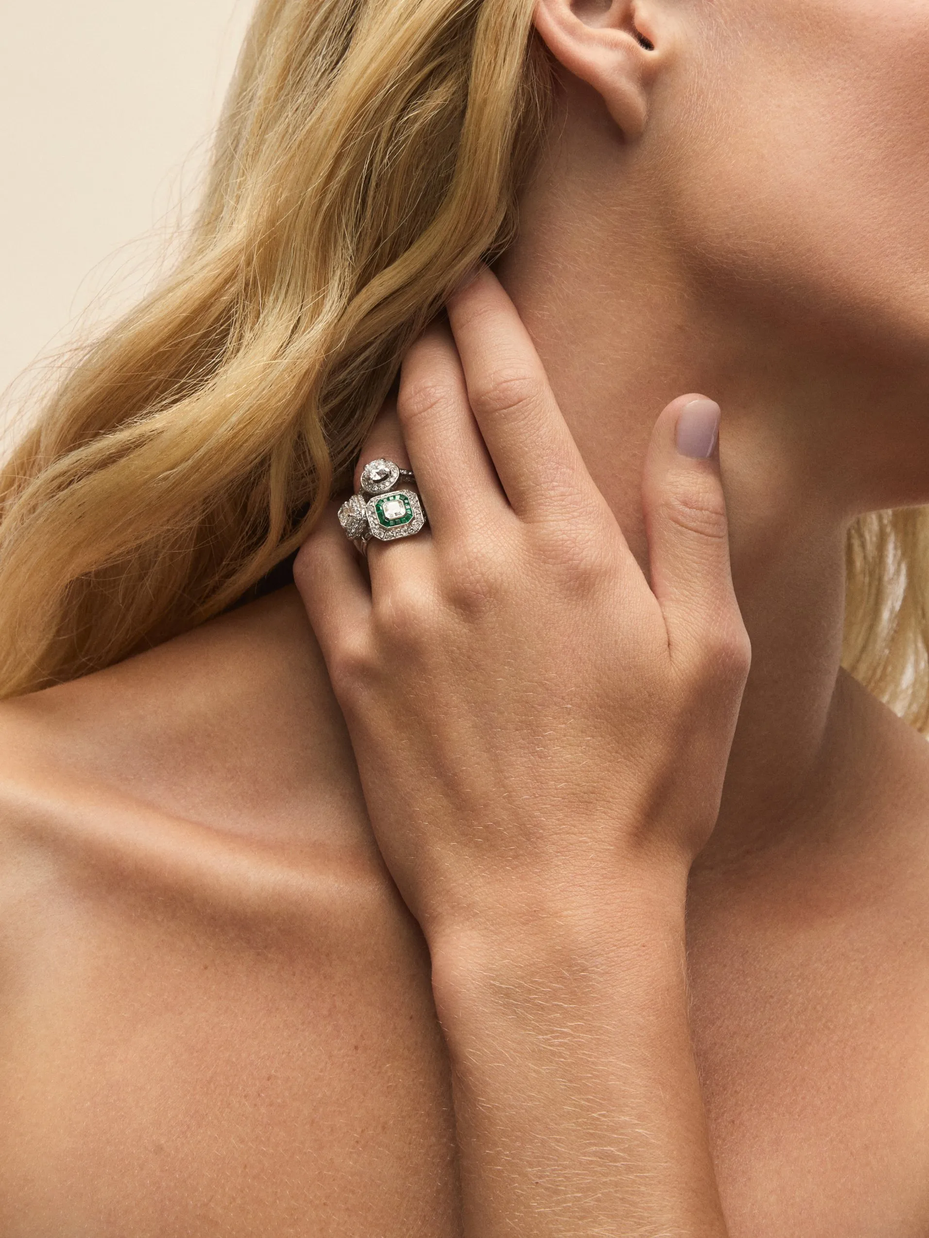 Close-up of woman wearing vintage diamond and emerald cluster ring, hand on neck, luxury jewellery detail.