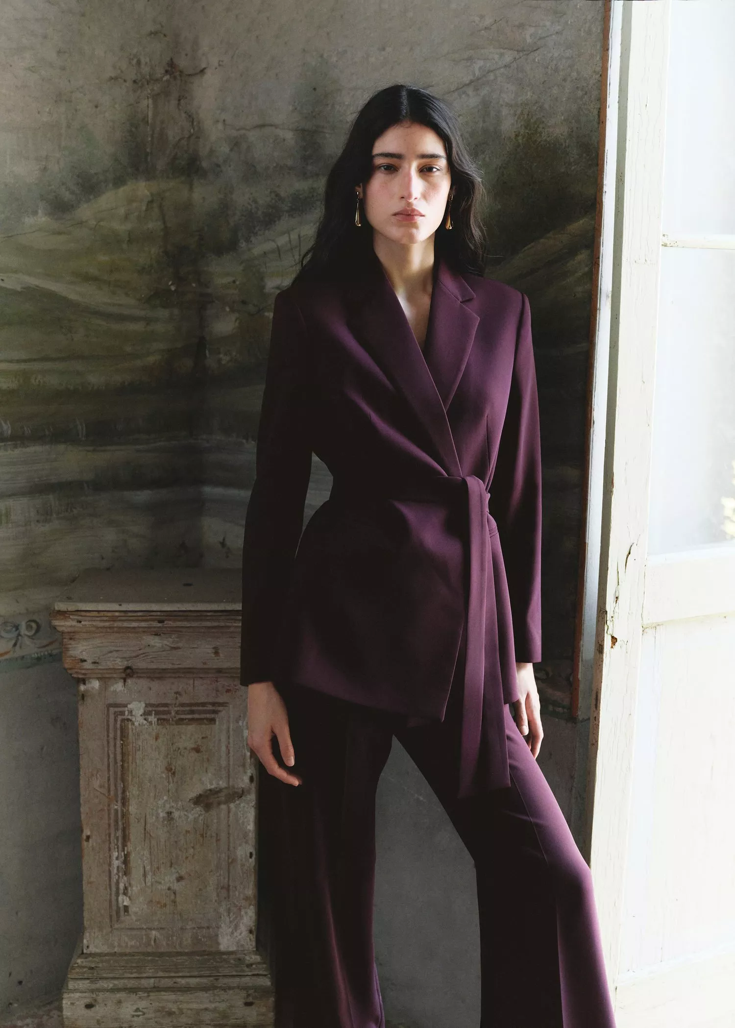 Fashion portrait of a woman in a plum wrap blazer and trousers, standing by a window in a rustic room.
