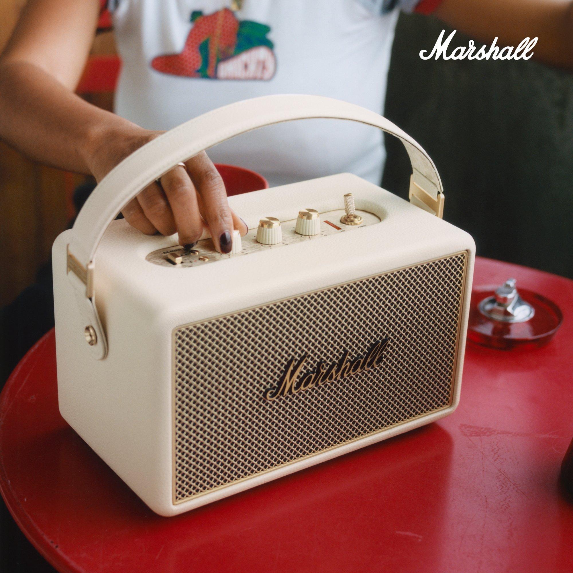 woman adjusts the knobs on a cream-colored Marshall portable Bluetooth speaker, placed on a red table.