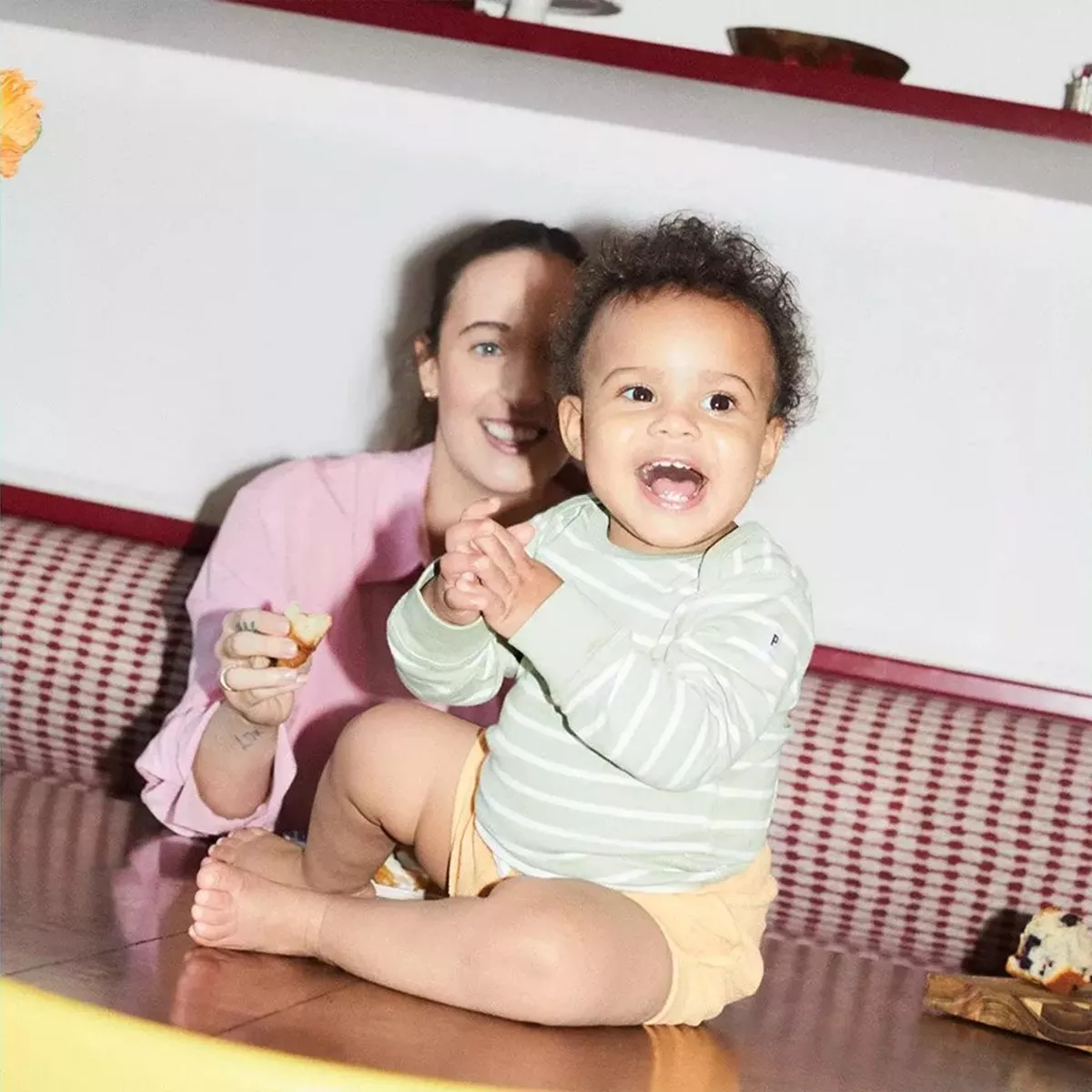 smiling baby with dark curly hair sitting barefoot on a table in the foreground, clapping its hands and looking at the camera, while a smiling adult in a pink shirt is blurred in the background