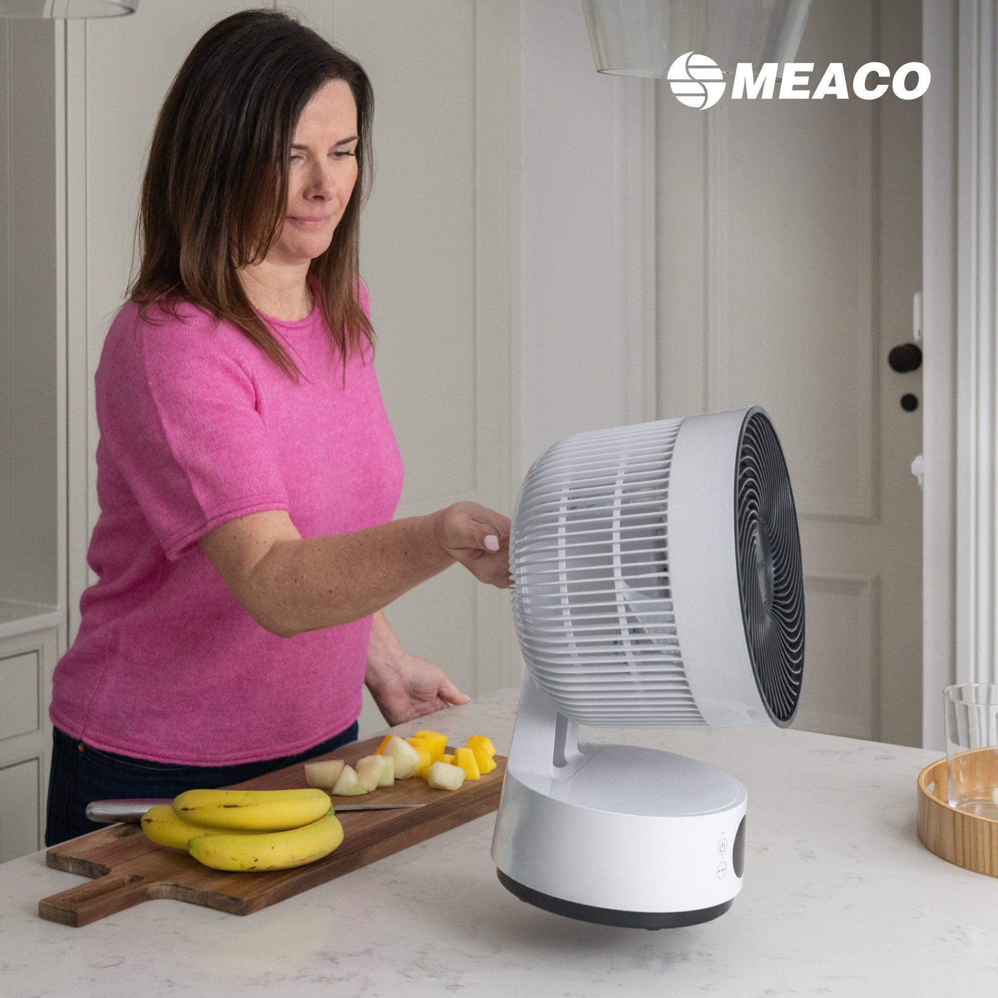 Woman in pink top adjusting a MEACO fan on a kitchen counter beside a cutting board with sliced fruit.