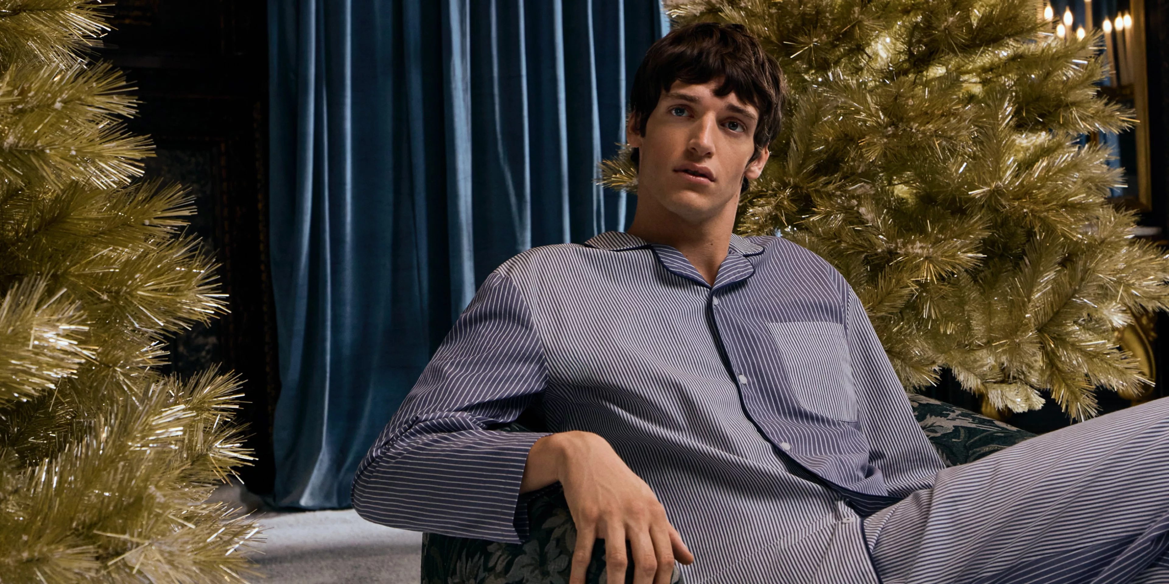 Man wearing striped blued pyjamas sat on an armchair in front of a gold Christmas tree