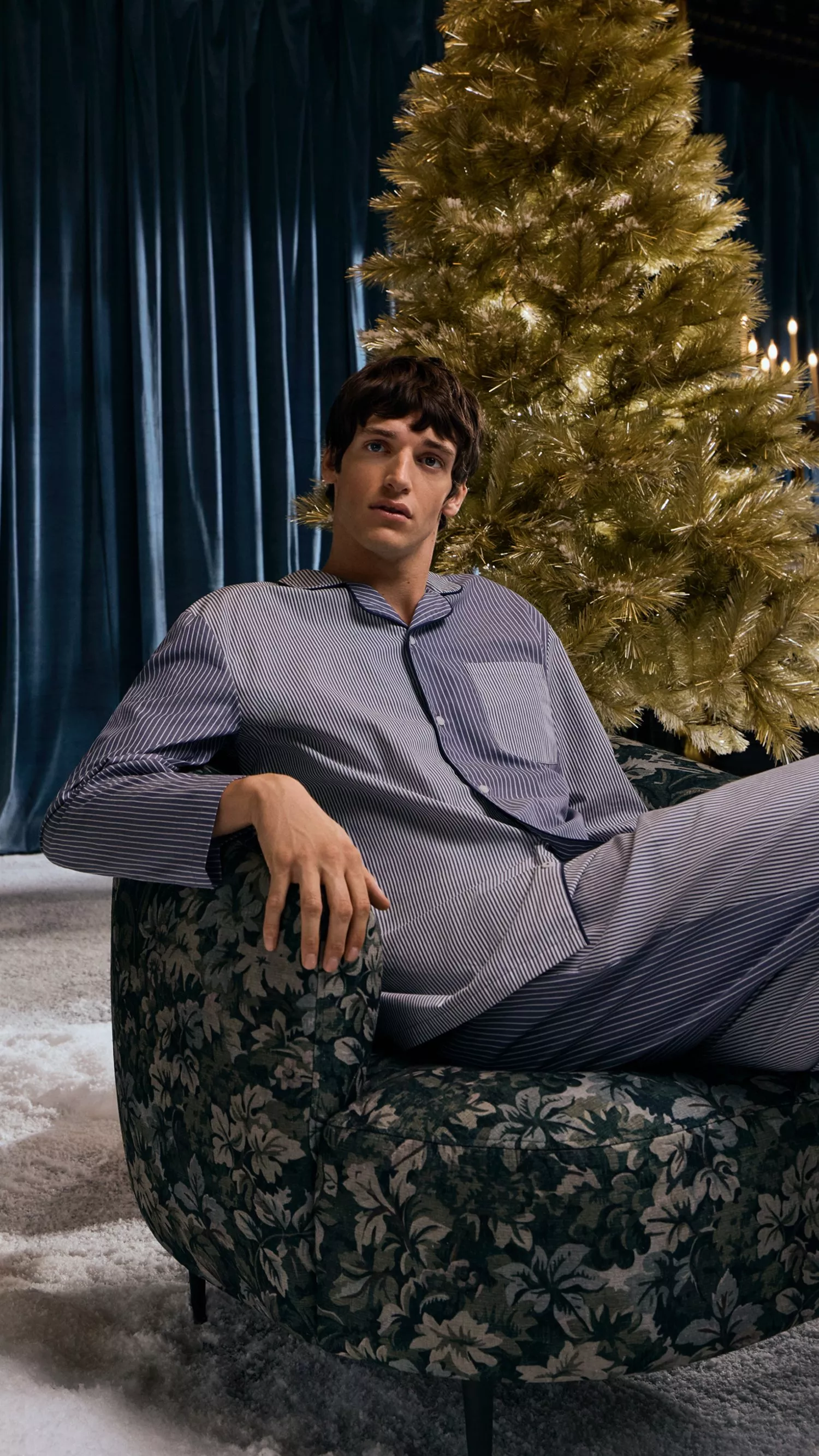 Man wearing striped blued pyjamas sat on an armchair in front of a gold Christmas tree