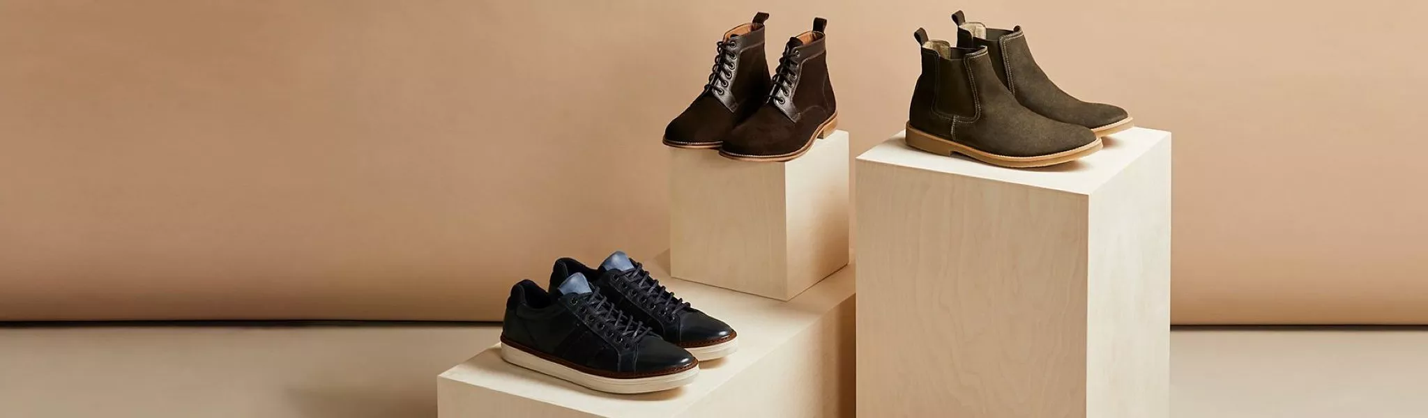 Men’s leather boots and trainers displayed on wooden plinths against a beige studio backdrop for footwear styling