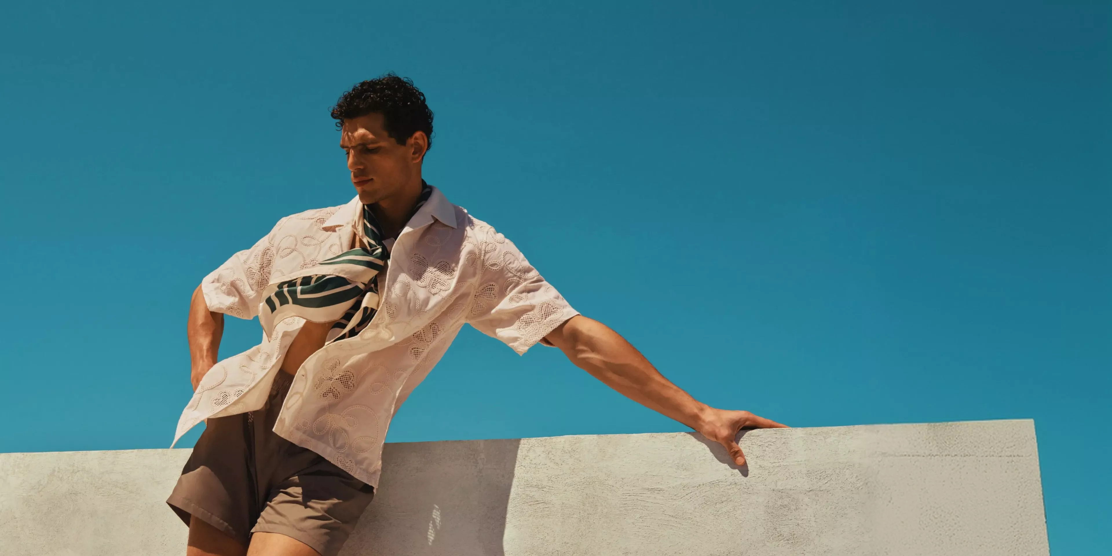 Man in summer outfit leaning on a white wall under clear blue sky, stylish menswear fashion photo outdoors