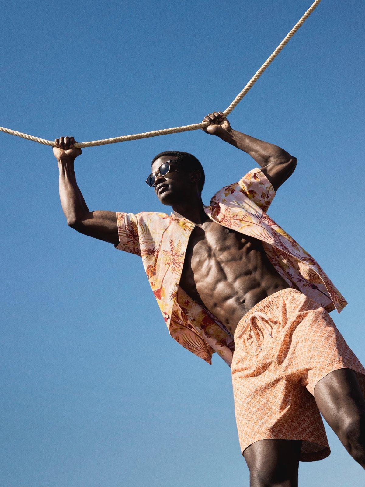 a model holding onto a rope with blue sky behind him, wearing a two piece shirt and shorts combo