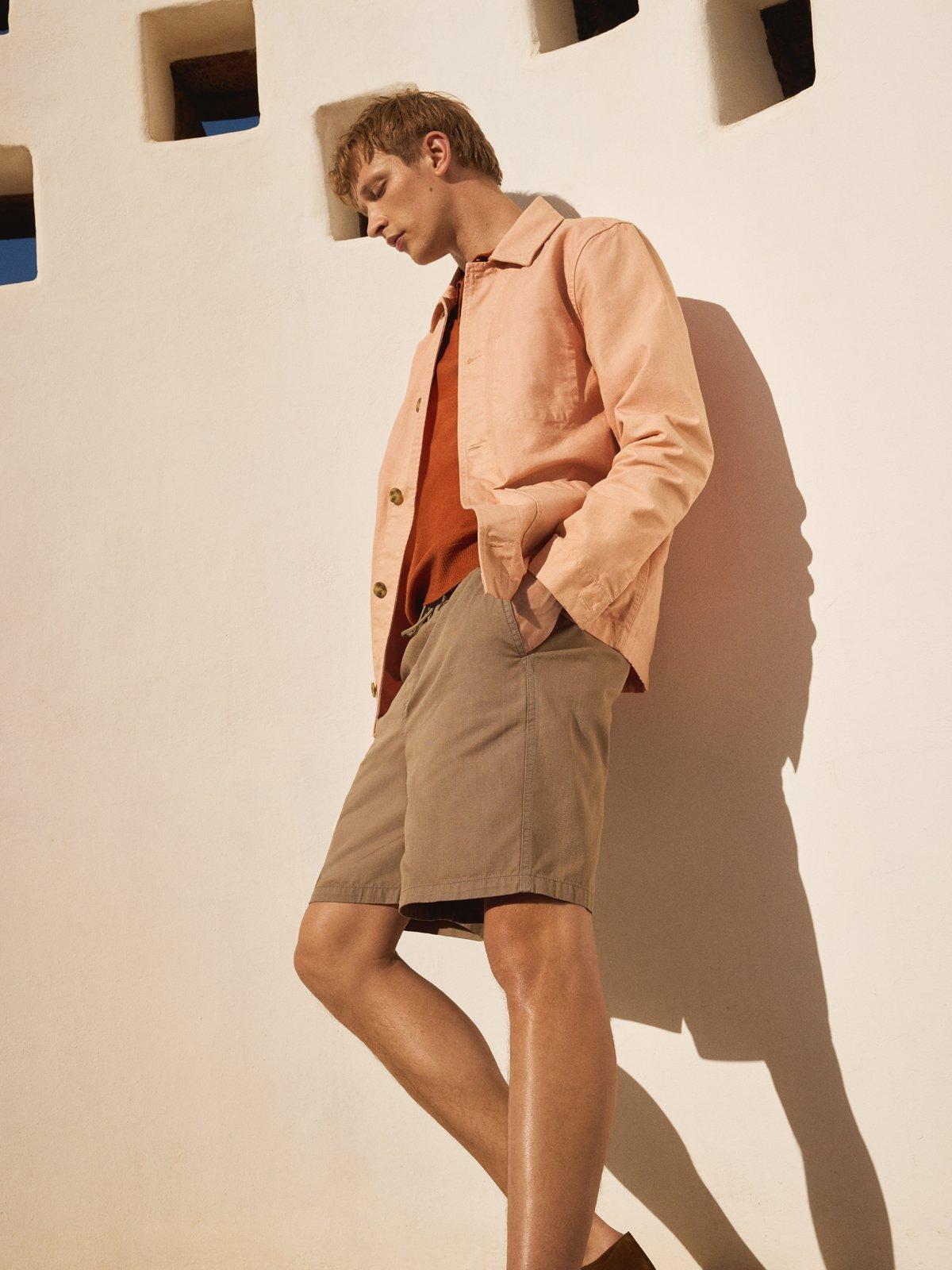 a model leaning against a wall in the sun wearing shorts and a shirt