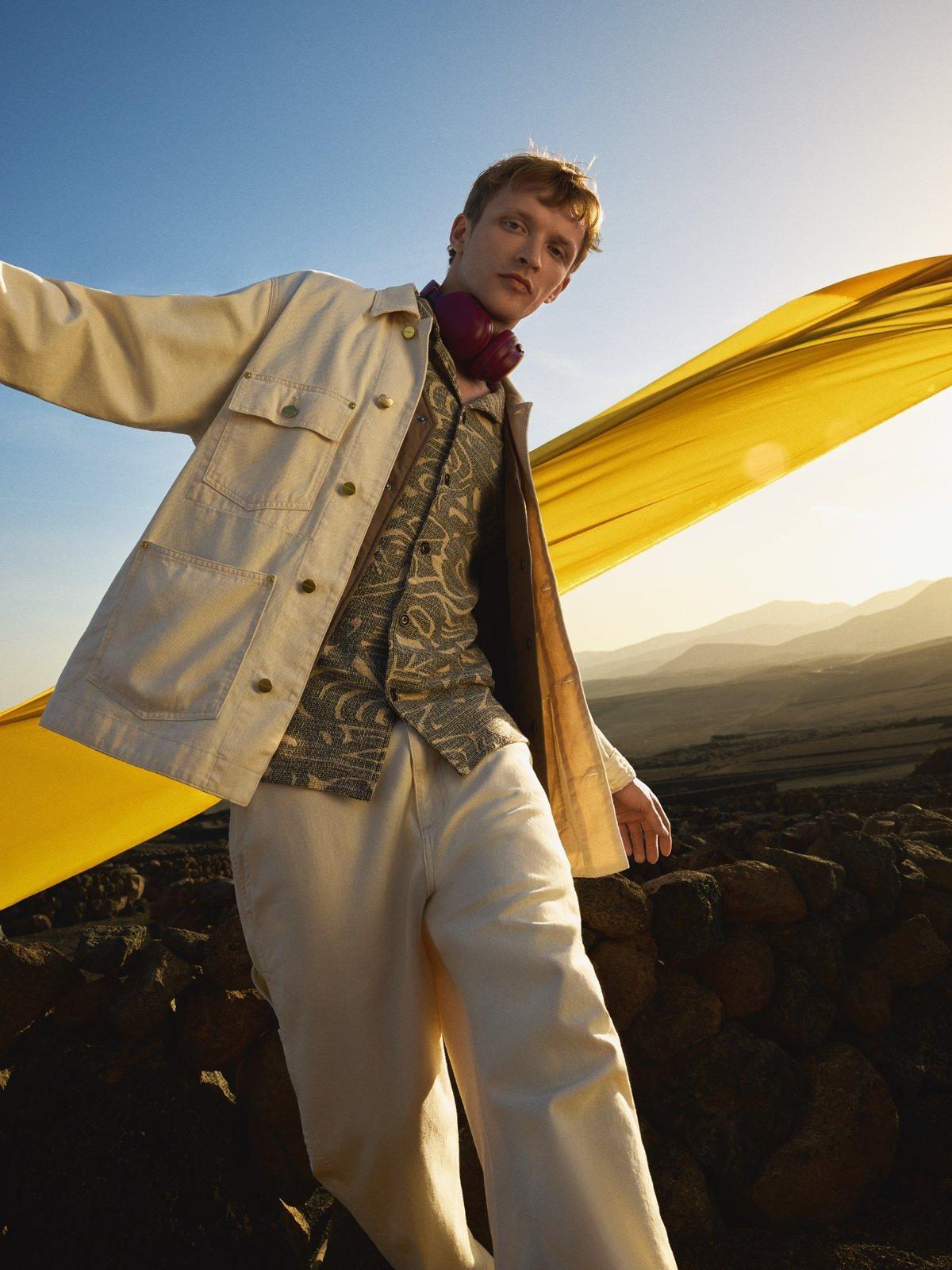 a model wearing a light beige jacket with a yellow flag and the sunset behind him