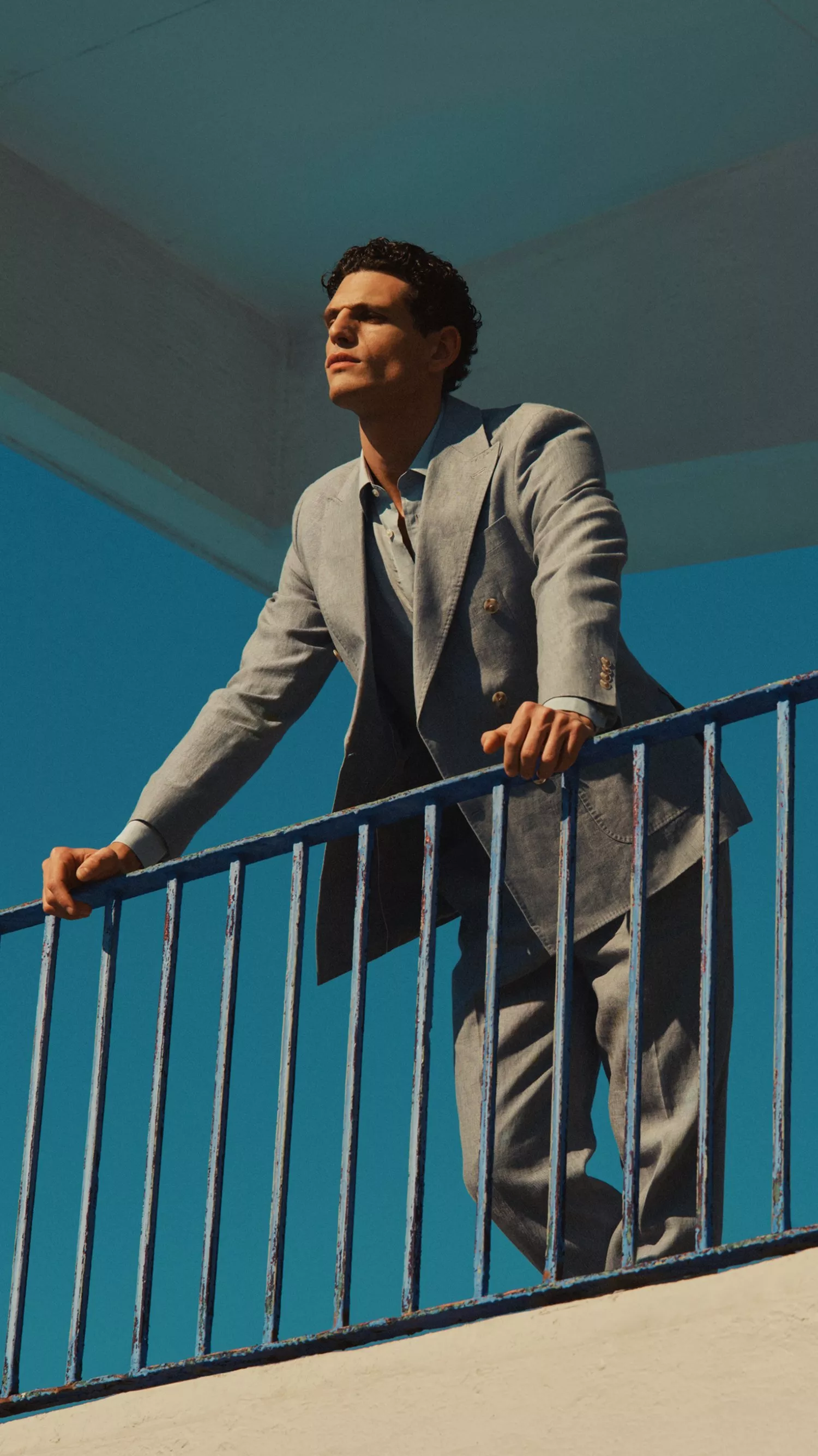 Stylish man in a light grey suit leaning on a blue railing beneath modern concrete architecture, blue sky backdrop