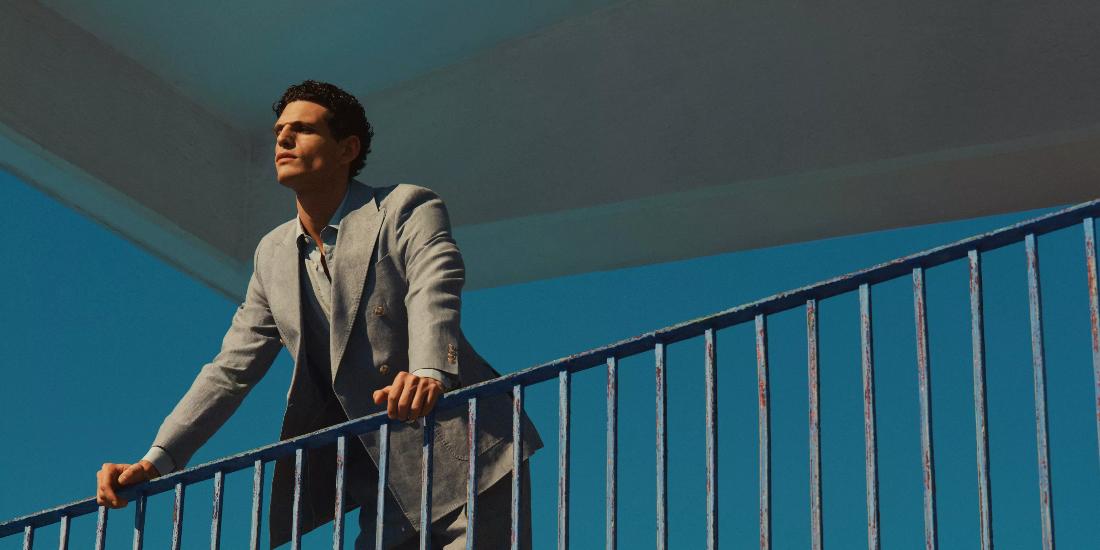 Man in light grey suit on outdoor stair railing, modern fashion portrait against bright blue sky and concrete canopy
