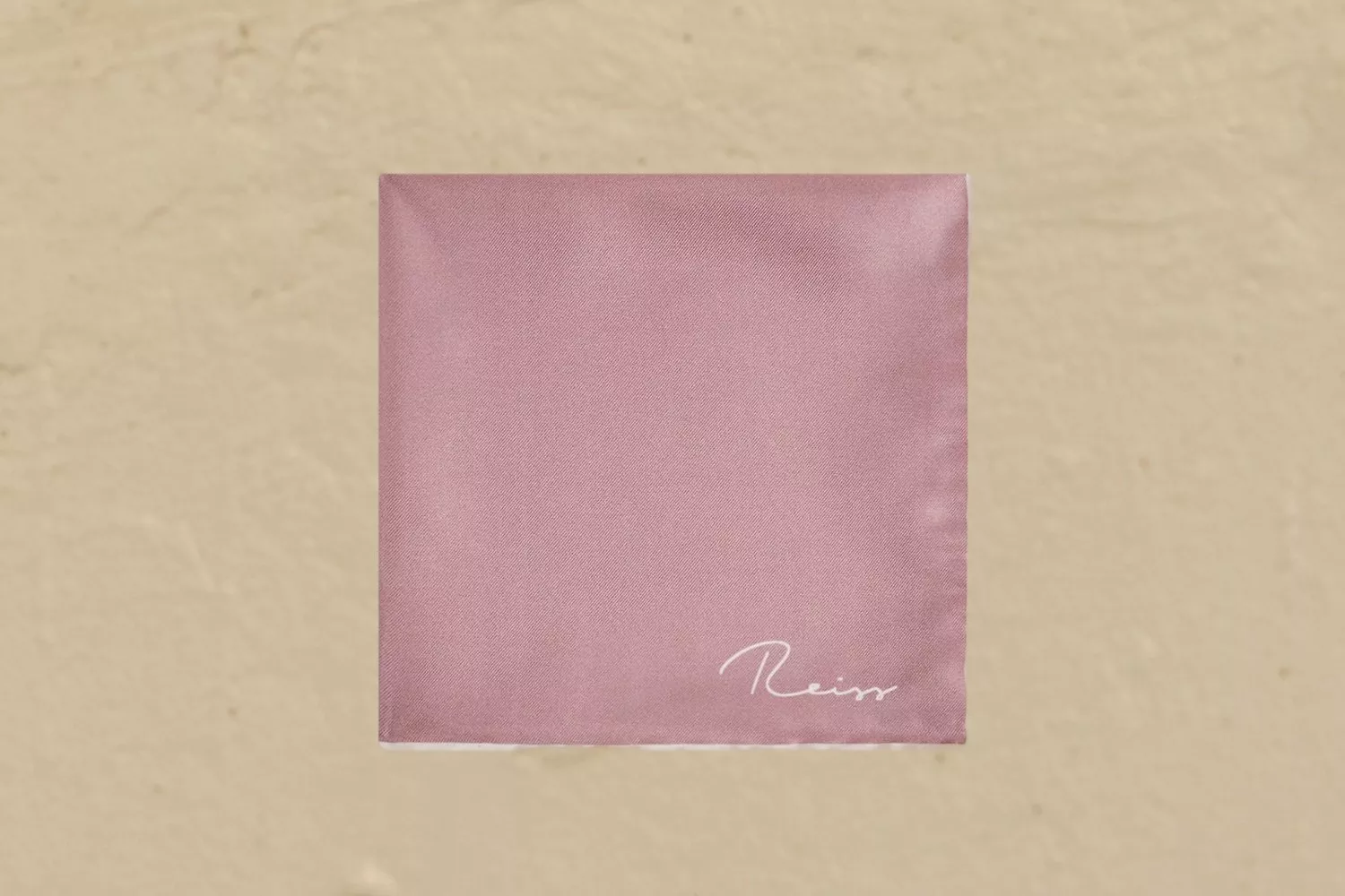 Pink fabric pocket square with white stitched logo, laid on beige background for menswear accessory styling