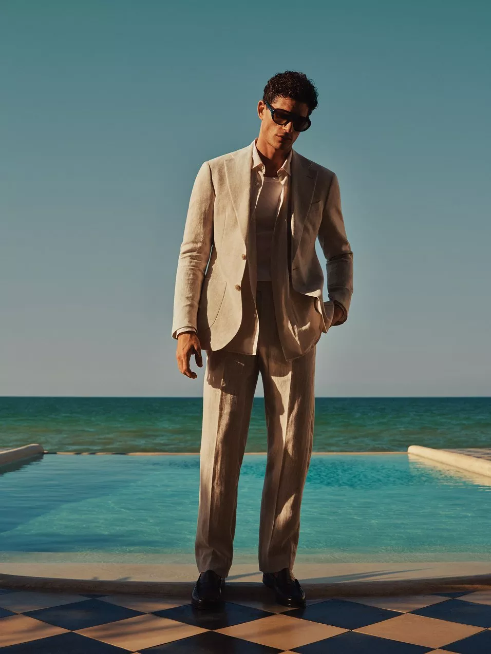 Man in beige linen suit and sunglasses by a pool with sea view