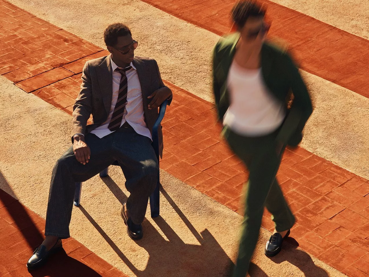 Man in suit sitting on chair outdoors on terracotta tiles, while blurred person in a green suit and loafers walks past