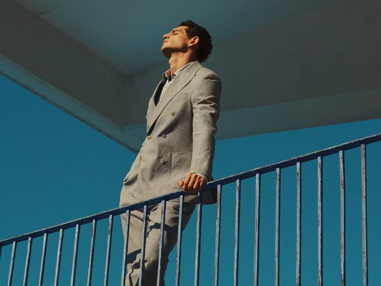 Man in light grey double-breasted suit leaning on balcony railing, against blue sky