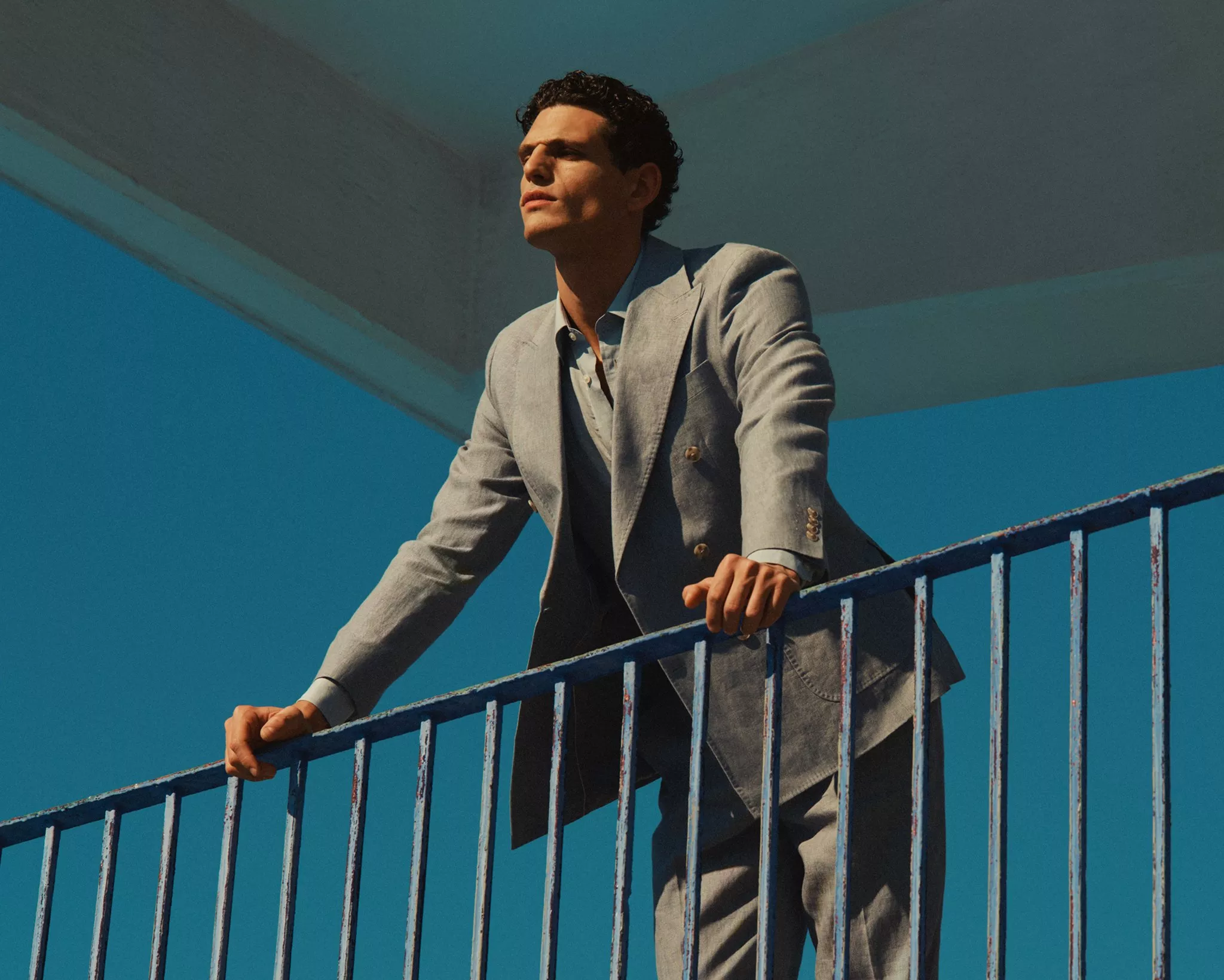 Man in light grey suit leaning on blue railing outdoors against a blue sky background