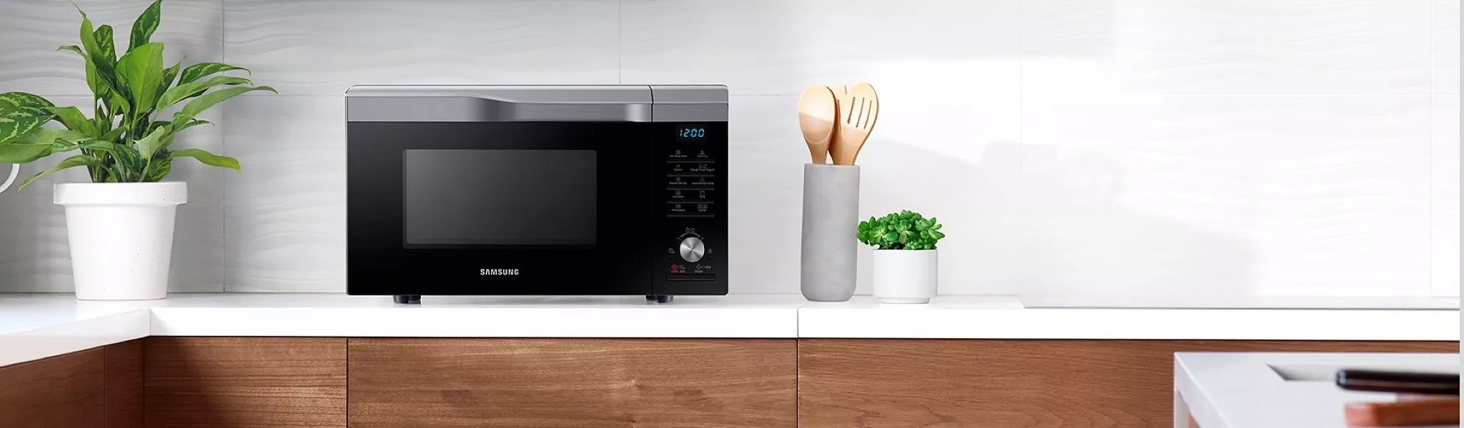Black Samsung microwave on modern kitchen worktop with potted plants and utensil holder, clean white tiles