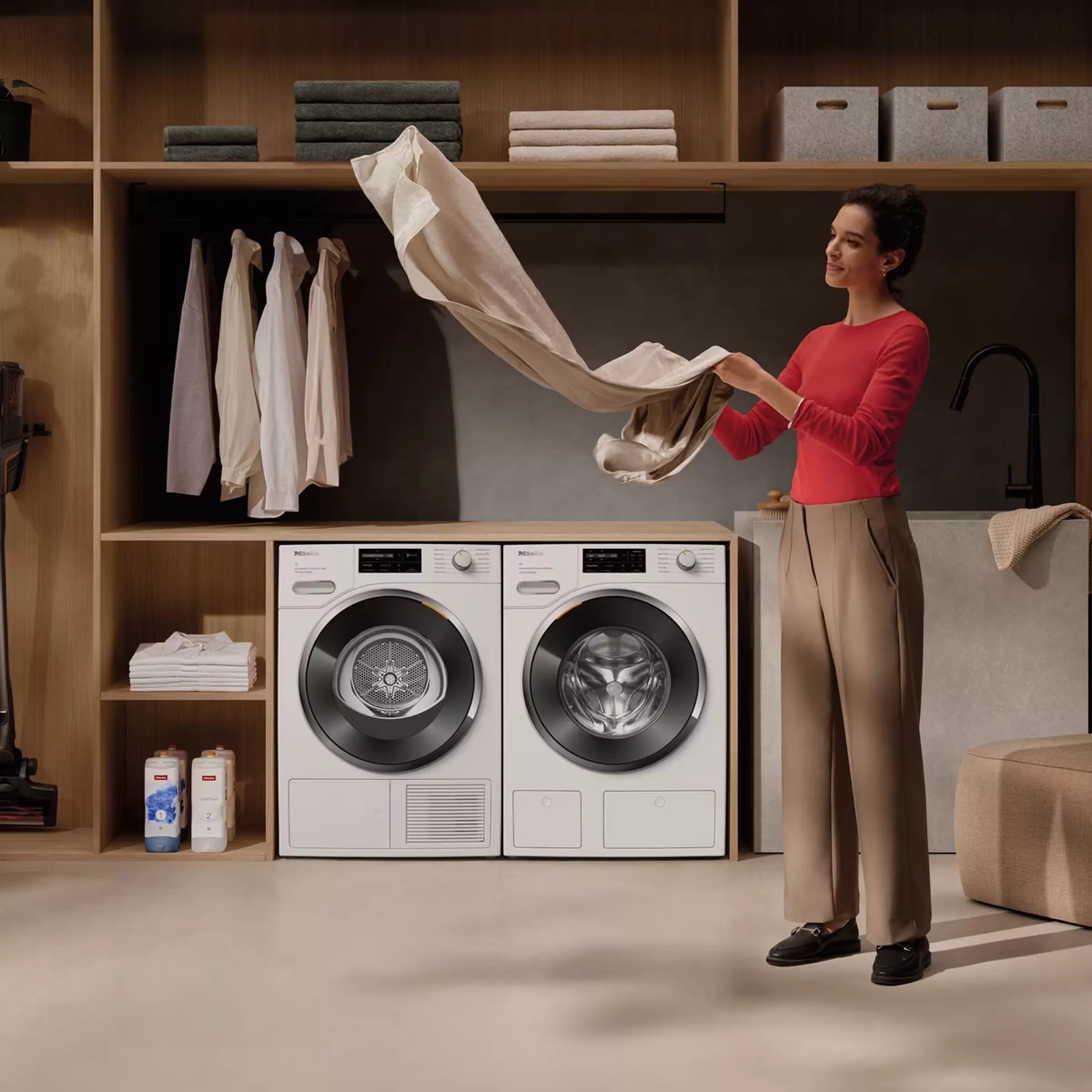 A woman in a red top folds laundry beside modern washing and drying machines in a tidy utility room.