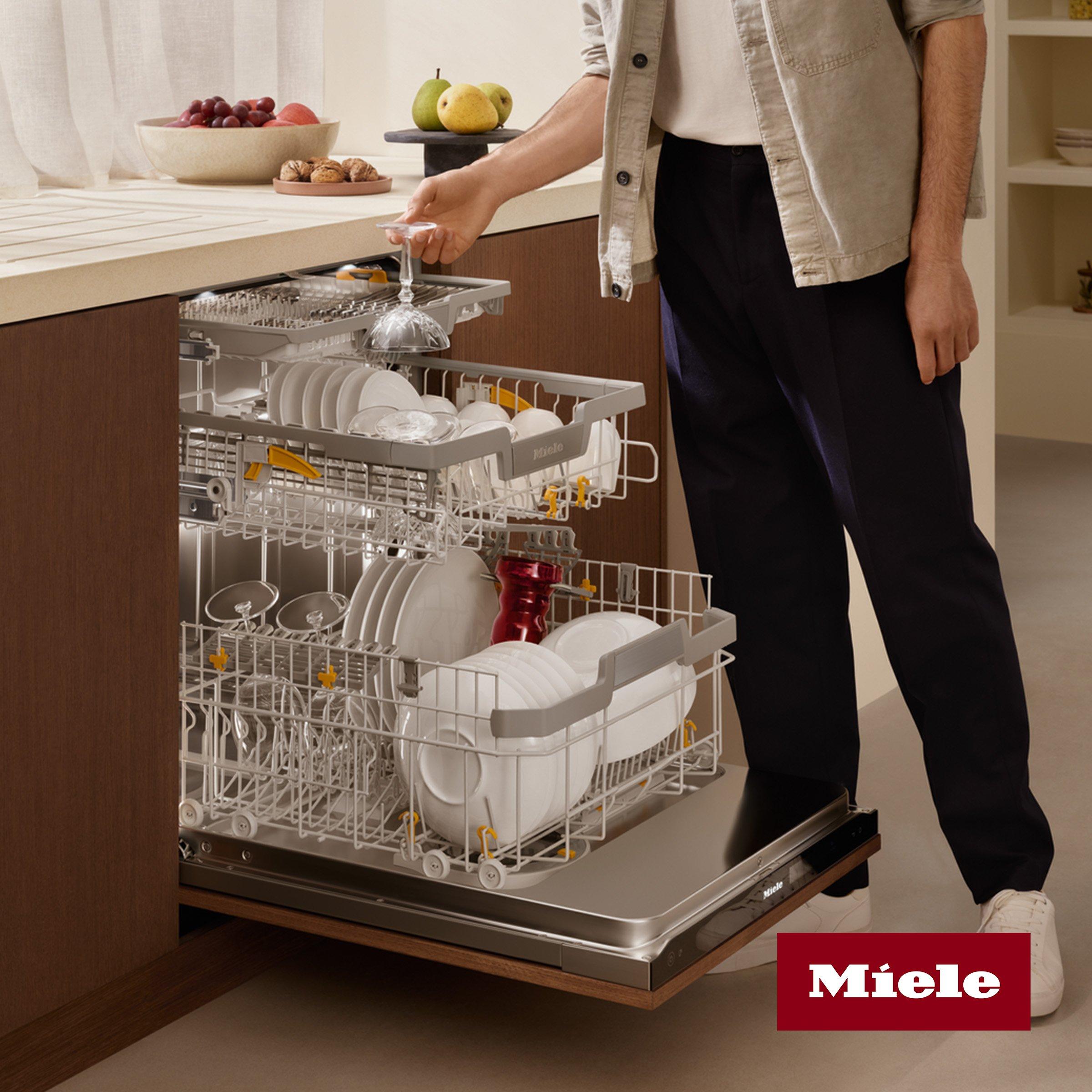 A person unloads clean dishes from a fully loaded Miele dishwasher in a modern kitchen.