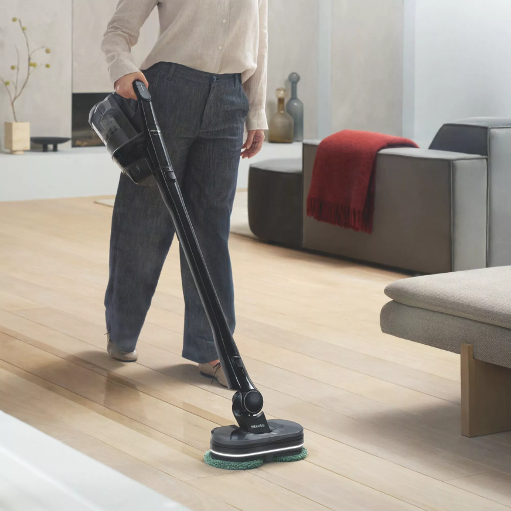 Person using cordless floor cleaner to scrub wooden floor in modern living room with sofa and red throw