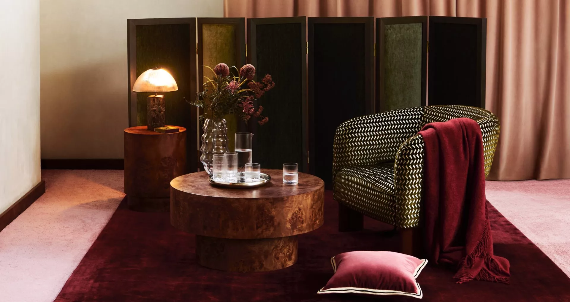 Cosy modern lounge with patterned armchair, wood coffee table, warm lamp, flowers, and burgundy textiles