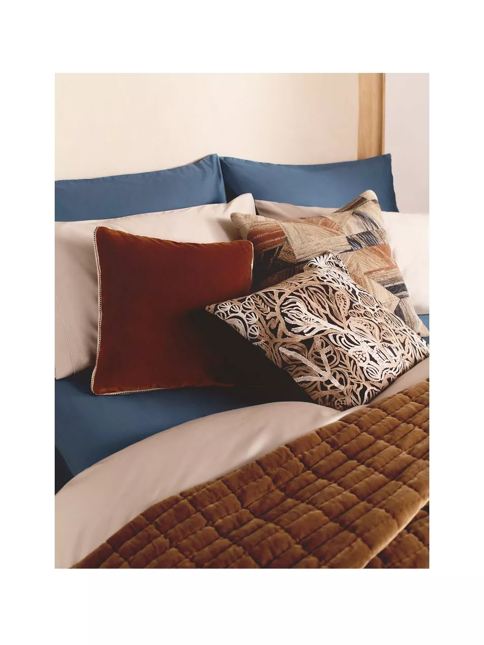 Luxurious bedding ensemble featuring rich, textured pillows and a soft quilt in warm earth tones and deep blue accents.