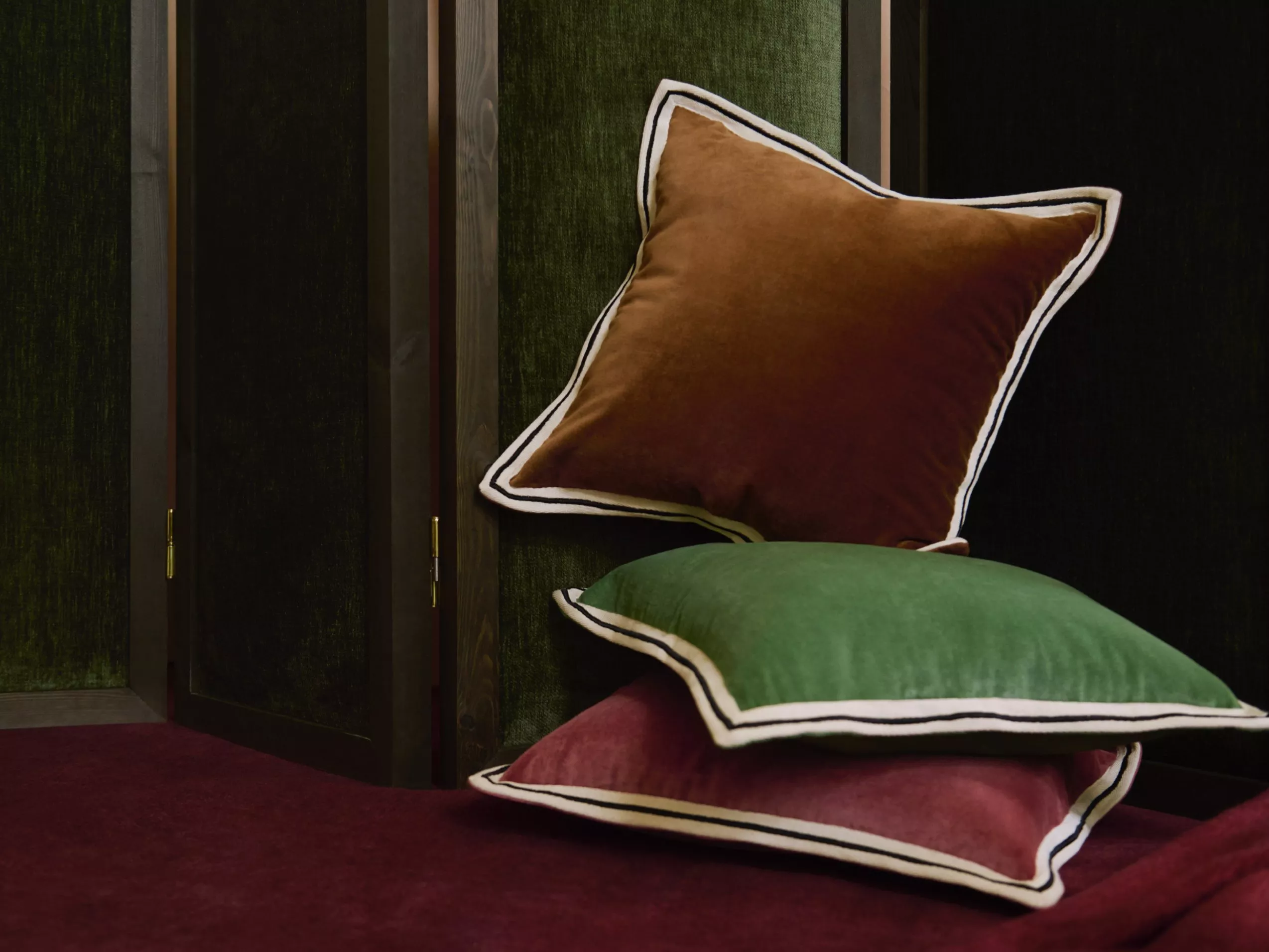 Stacked velvet cushions in green, brown and burgundy with contrast piping on a sofa by dark wooden panels