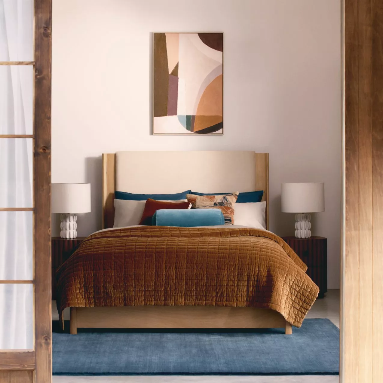 Modern bedroom with a beige headboard, rust-coloured quilt, and vibrant cushions. Abstract art hangs above the bed.