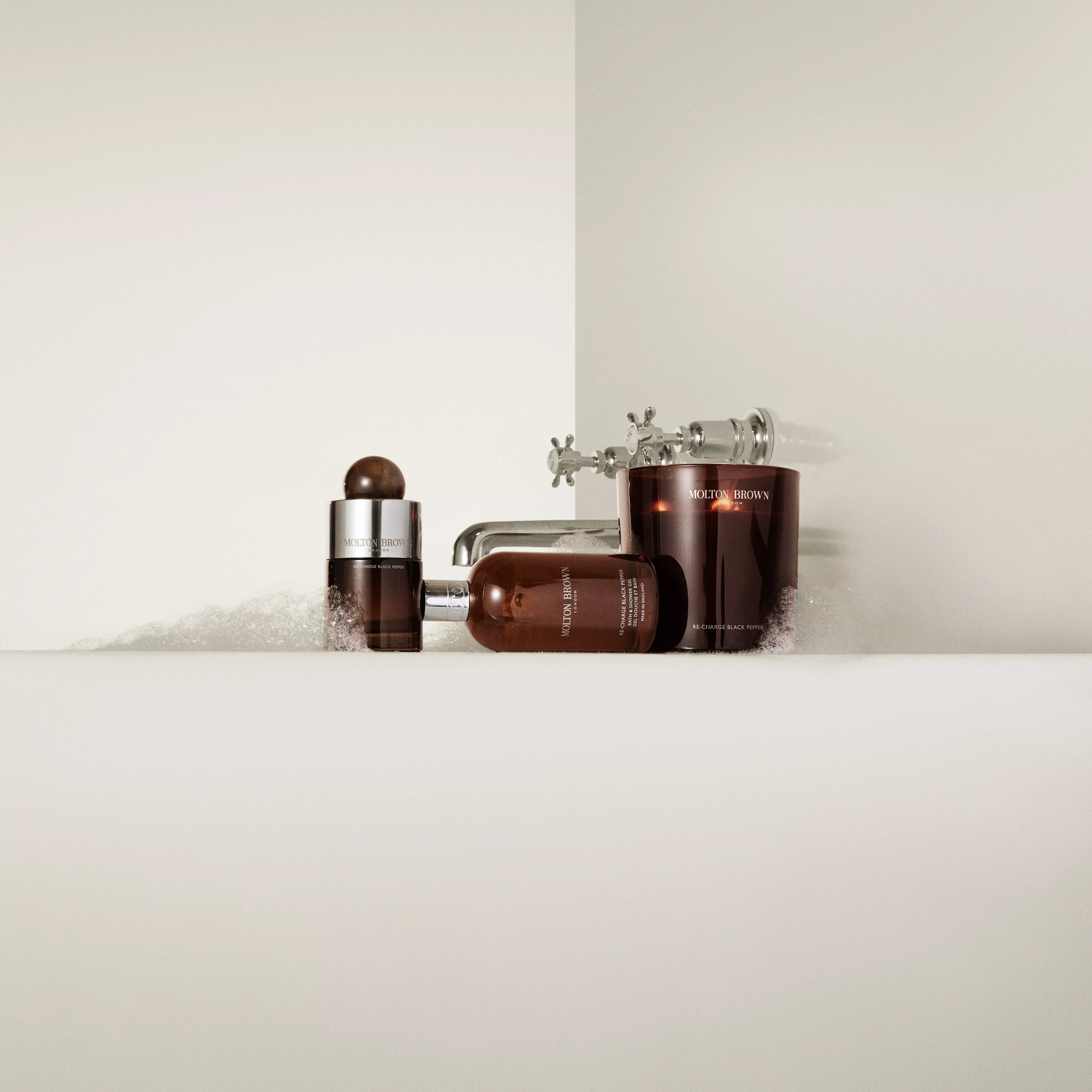 Molton Brown Re-charge Black Pepper bath and body products beside a chrome tap with bubbles in a white bath.