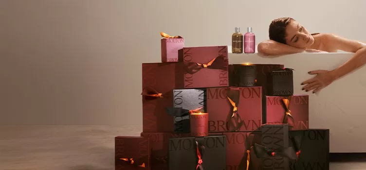 A brand banner showing a woman relaxing in a bathtub surrounded by Molton Brown gift boxes, lit candles, and bottles of bath products.