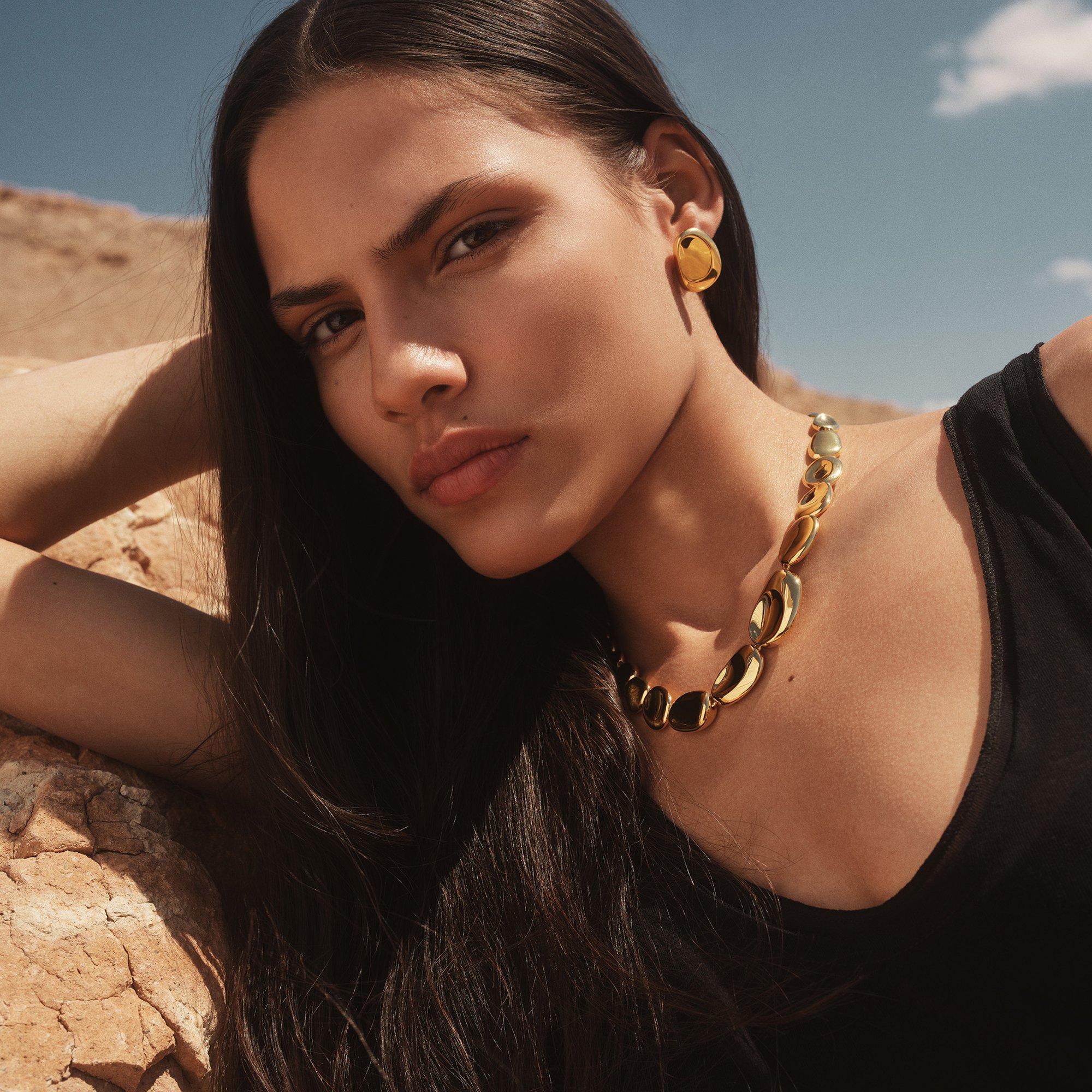 Woman wearing a chunky gold necklace and earrings poses outdoors against a rocky background.
