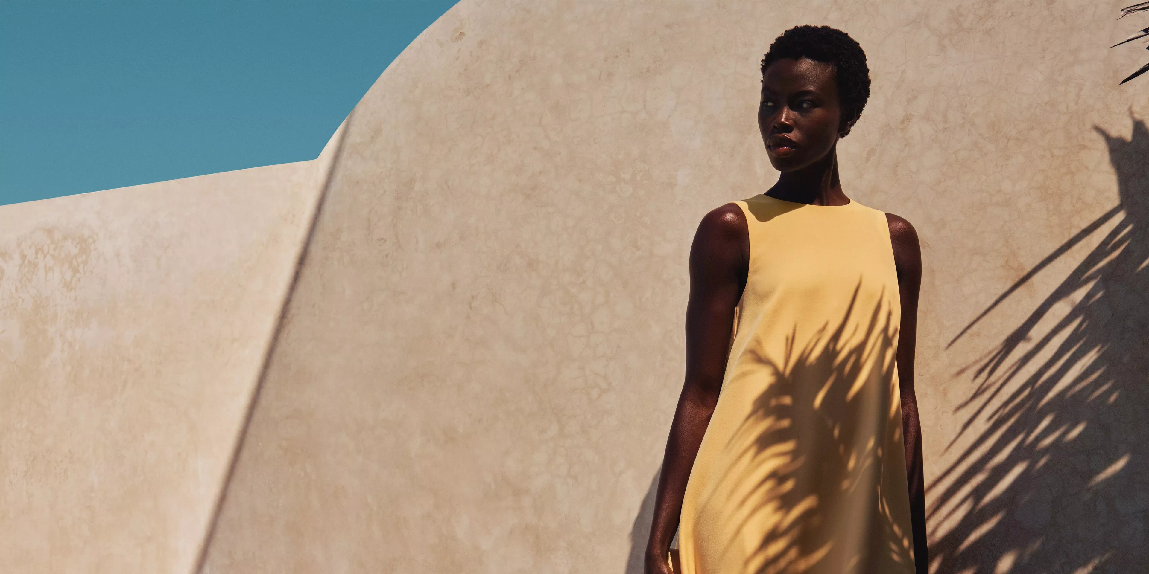 Fashion model in yellow sleeveless dress by sunlit stucco wall, palm shadows and blue sky backdrop