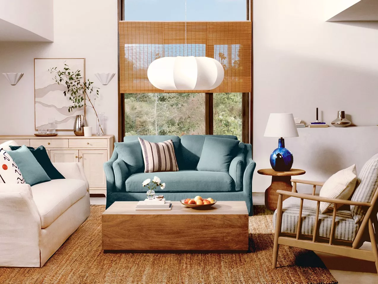 A light airy lounge with a tall ceiling and large window, a blue-green sofa, a white sofa, and wooden framed armchair.