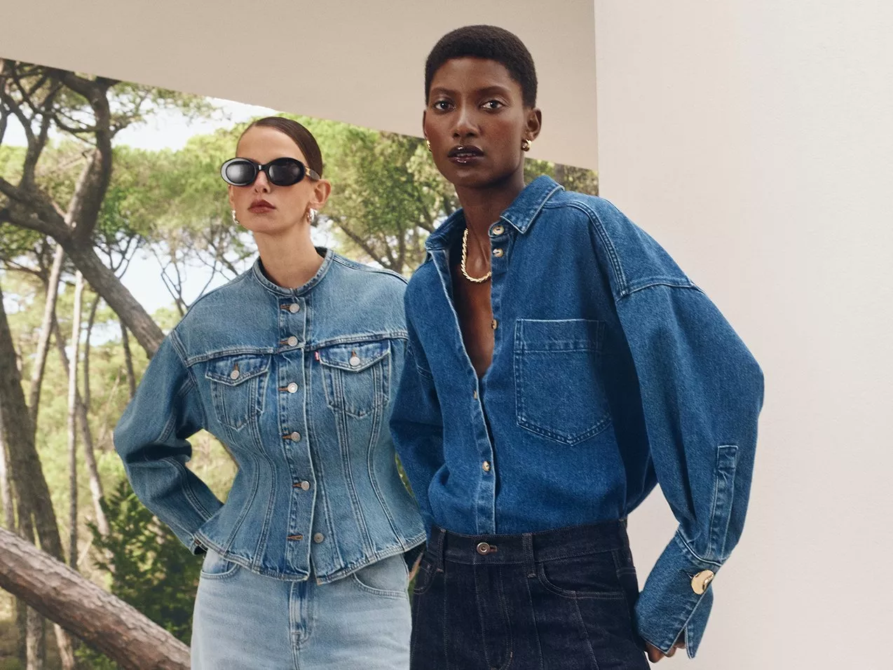 Two models in denim jackets next to a white outdoor structure with trees in the background.