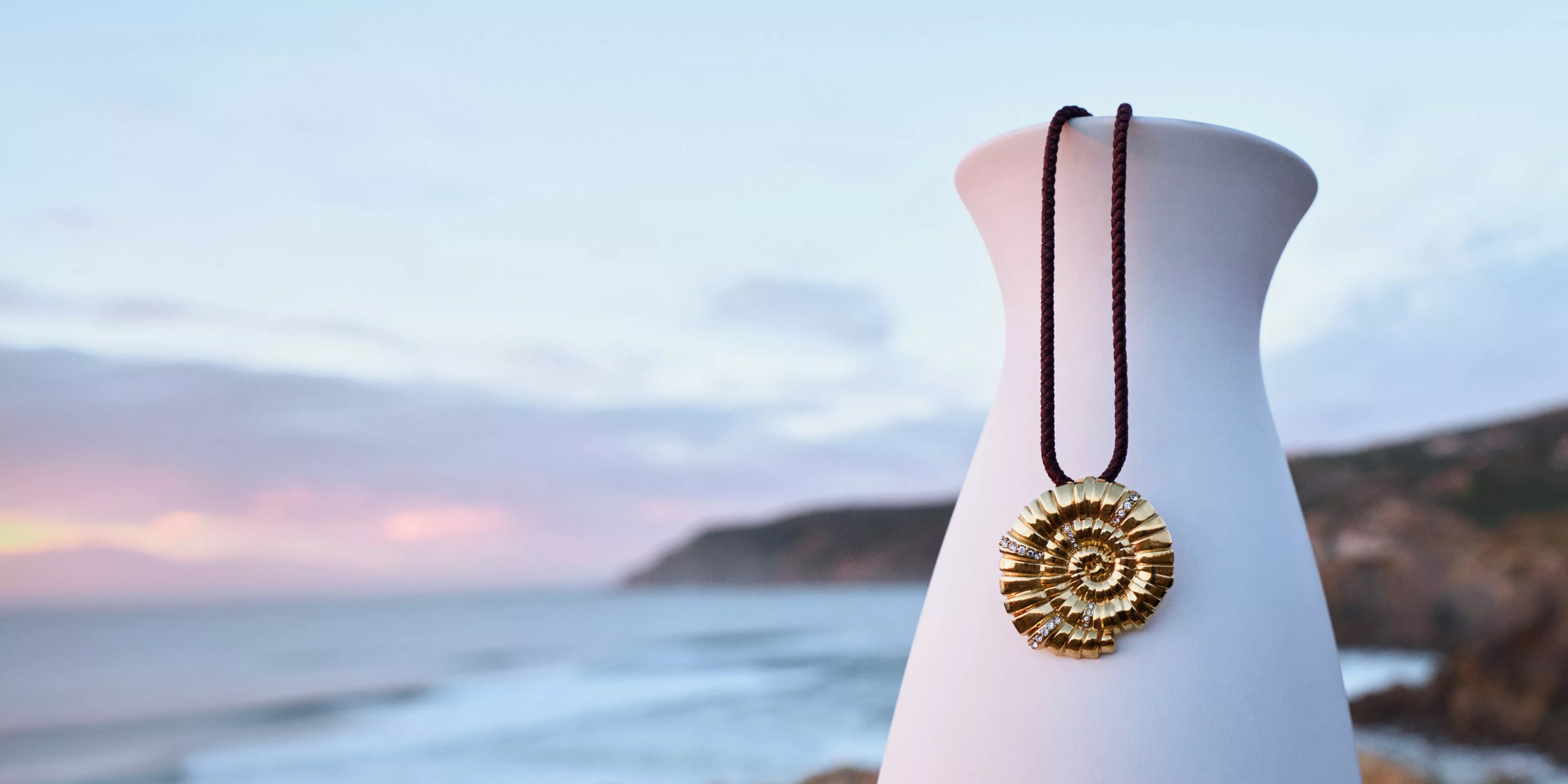 Gold ammonite pendant necklace displayed on white vase, with coastal background.