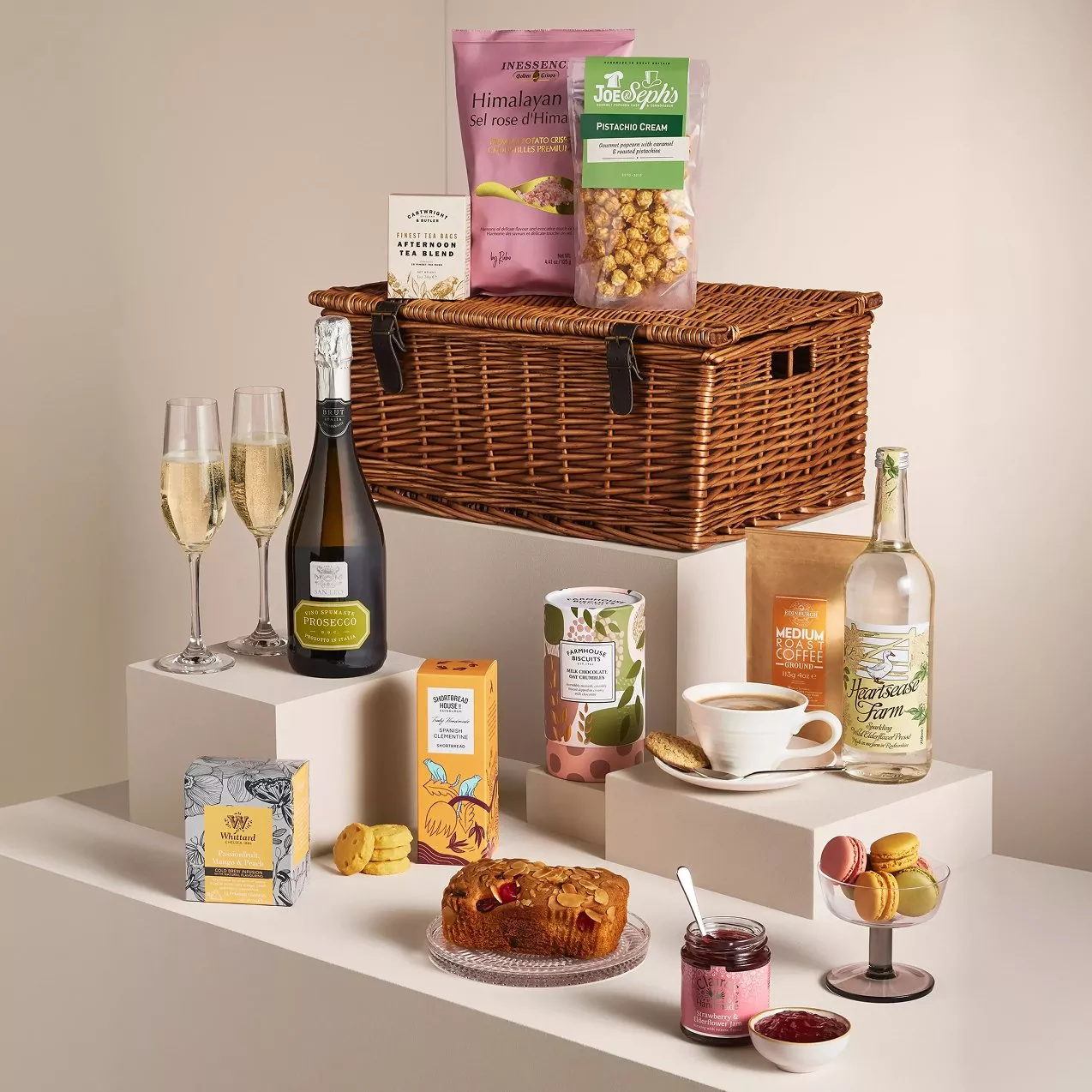 A luxurious gift hamper with Prosecco, gourmet snacks, and beverages, elegantly displayed on white pedestals.